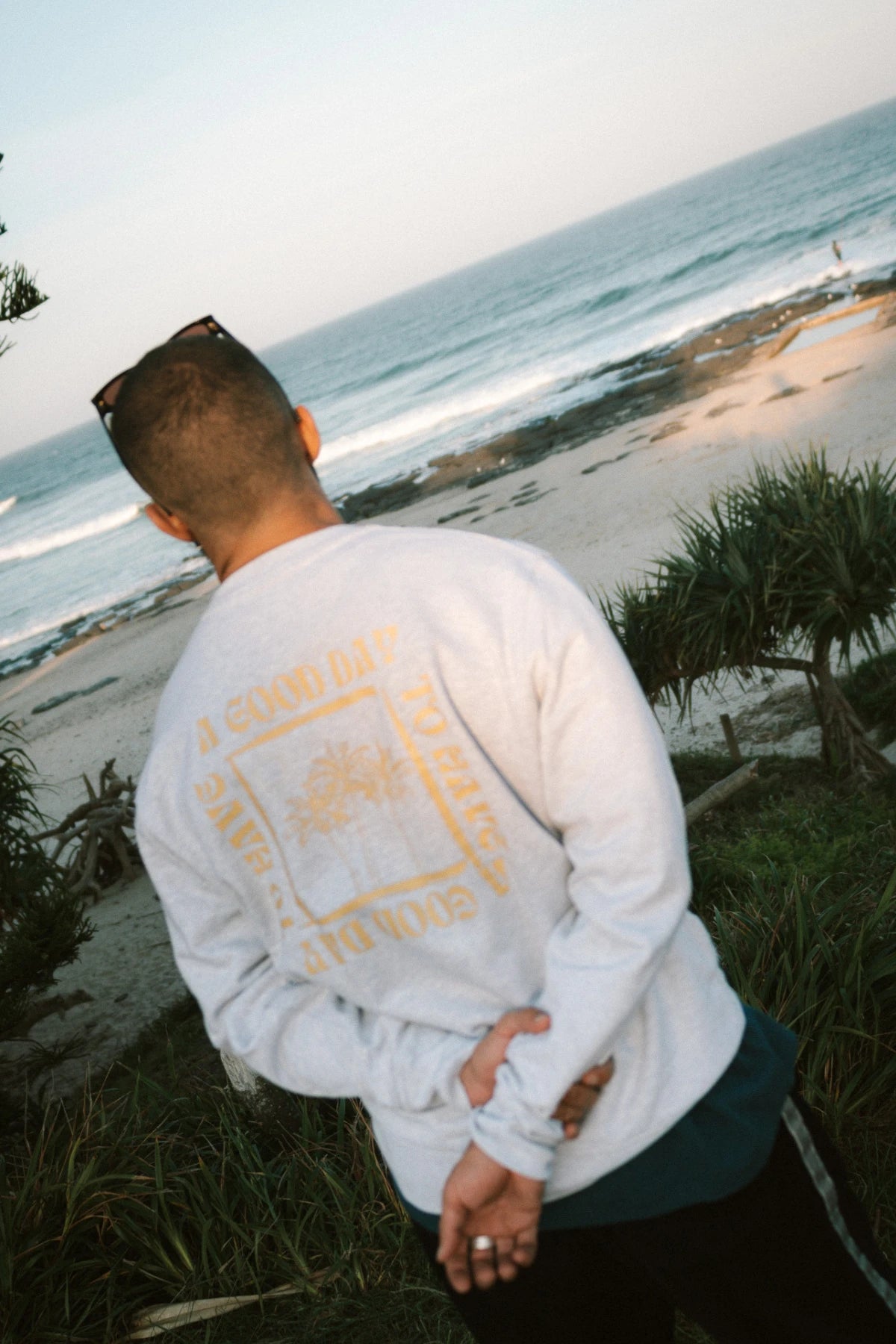 Rückansicht eines Mannes im A Good Day Sweater Cool Grey von The Sunkissed Rebels mit Blick auf das Meer – entspannter Surf-Vibe am Strand.