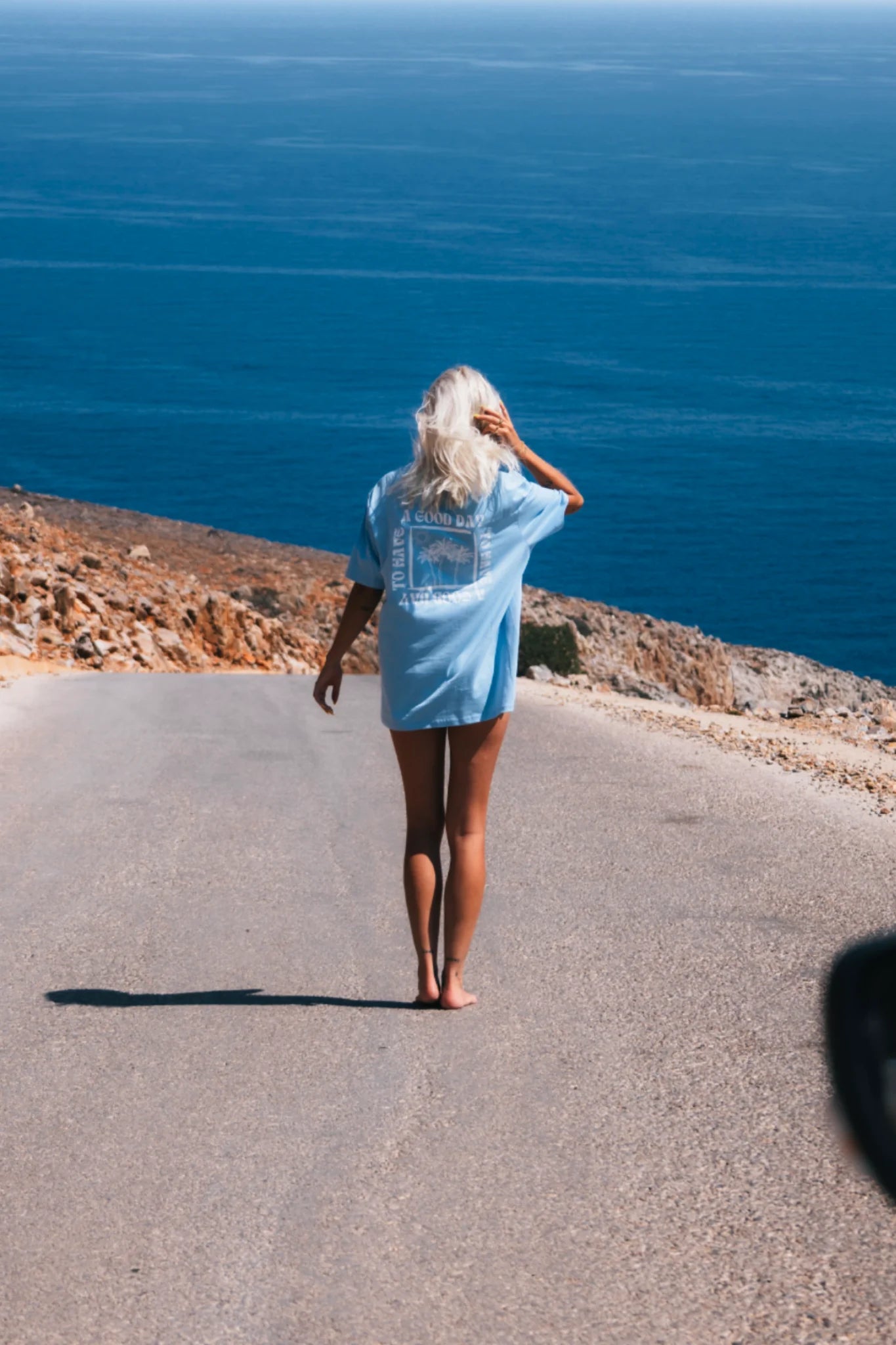 Frau läuft barfuß an Küstenstraße mit blauem A Good Day T-Shirt – Sunkissed Rebels Blue Soul Shirt am Meer