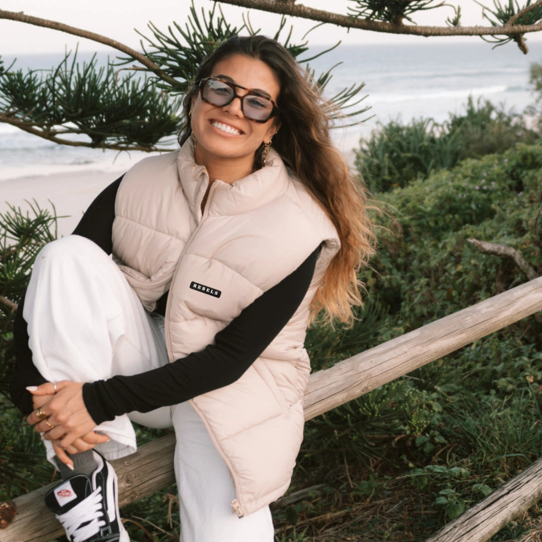 Frau mit beiger Pufferweste und Sonnenbrille lacht fröhlich am Meer, im Hintergrund grüne Pflanzen und Wellen.
