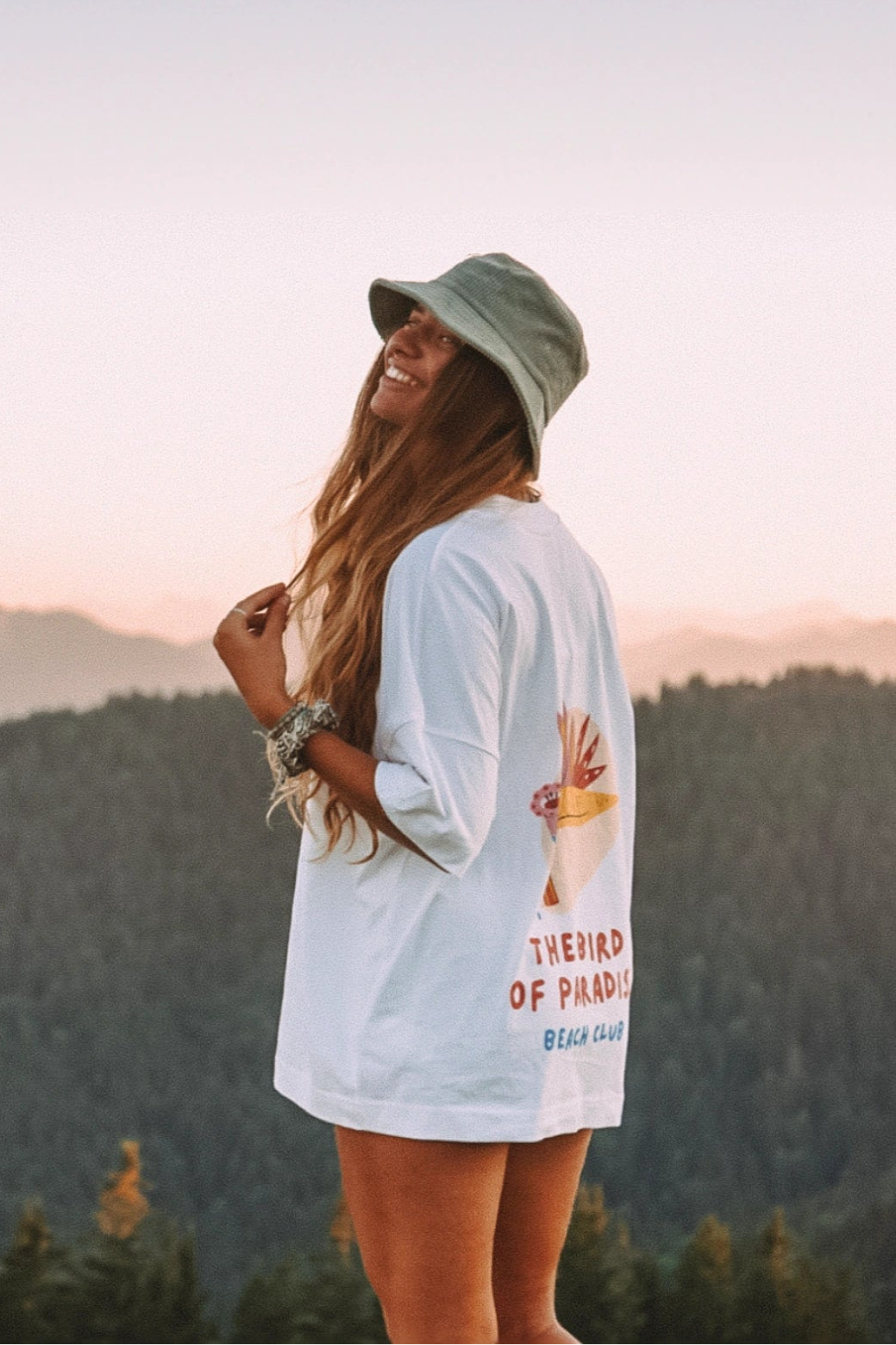 Seitliche Lifestyle Aufnahme einer Frau im weißen Bird of Paradise Boxy Oversized T Shirt bei Sonnenuntergang in den Bergen