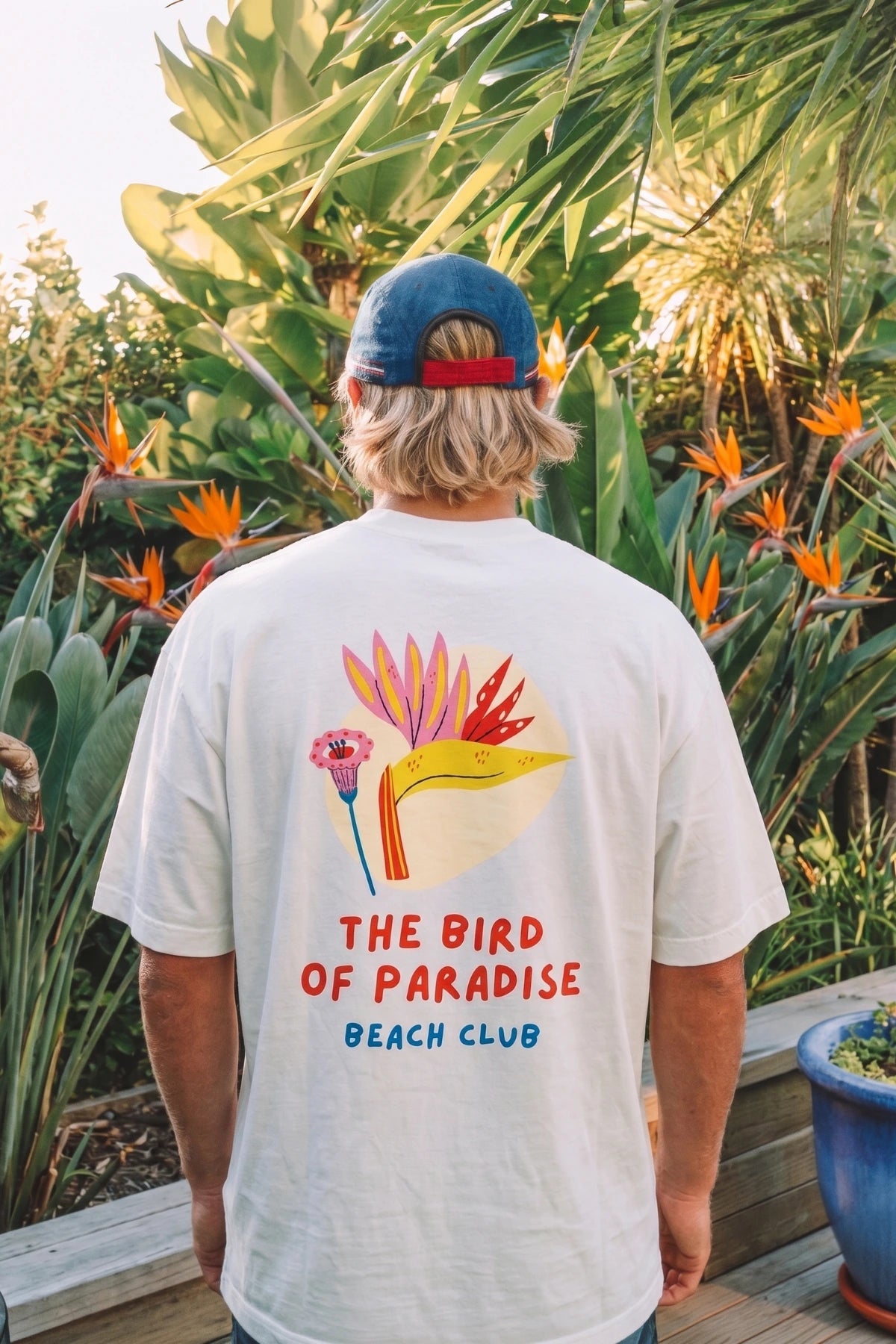 Lifestyle Aufnahme des weißen Bird of Paradise Boxy Oversized T-Shirts in tropischer Palmenkulisse mit Beach Club Design 