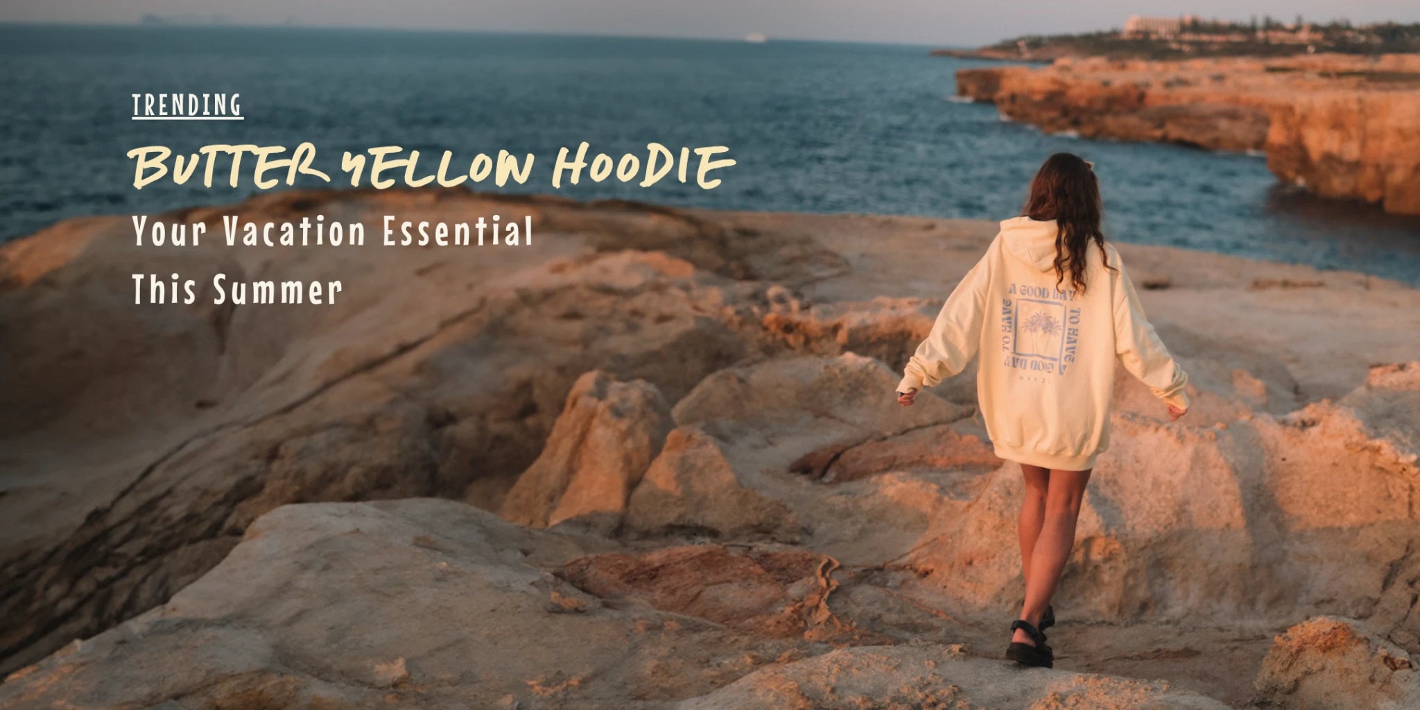 Woman in butter yellow hoodie walking on coastal rocks during golden hour – summer beachwear trend