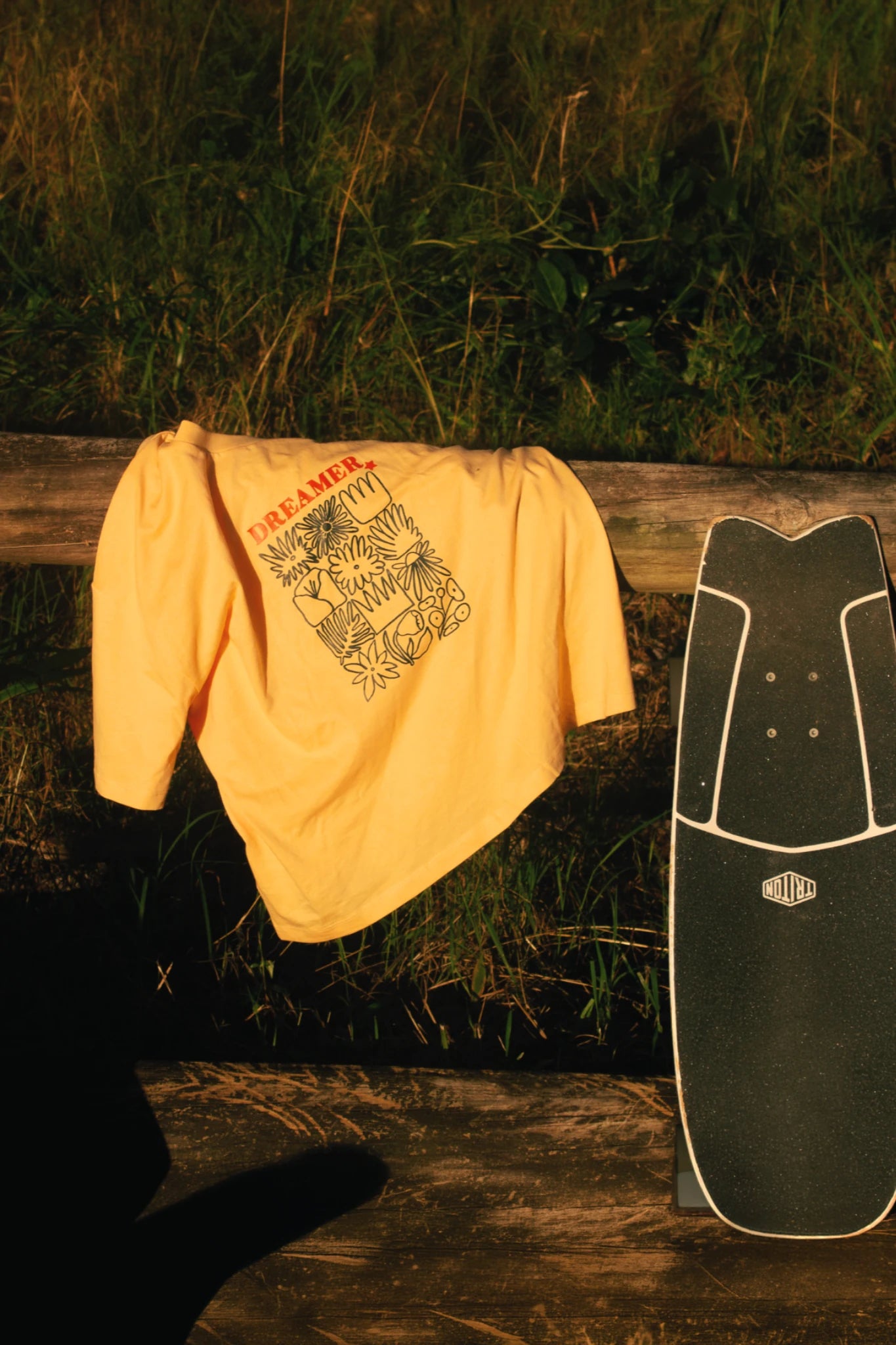 Papayafarbenes Dreamer T-Shirt mit großem Backprint liegt über Holzgeländer neben Skateboard im warmen Sonnenlicht