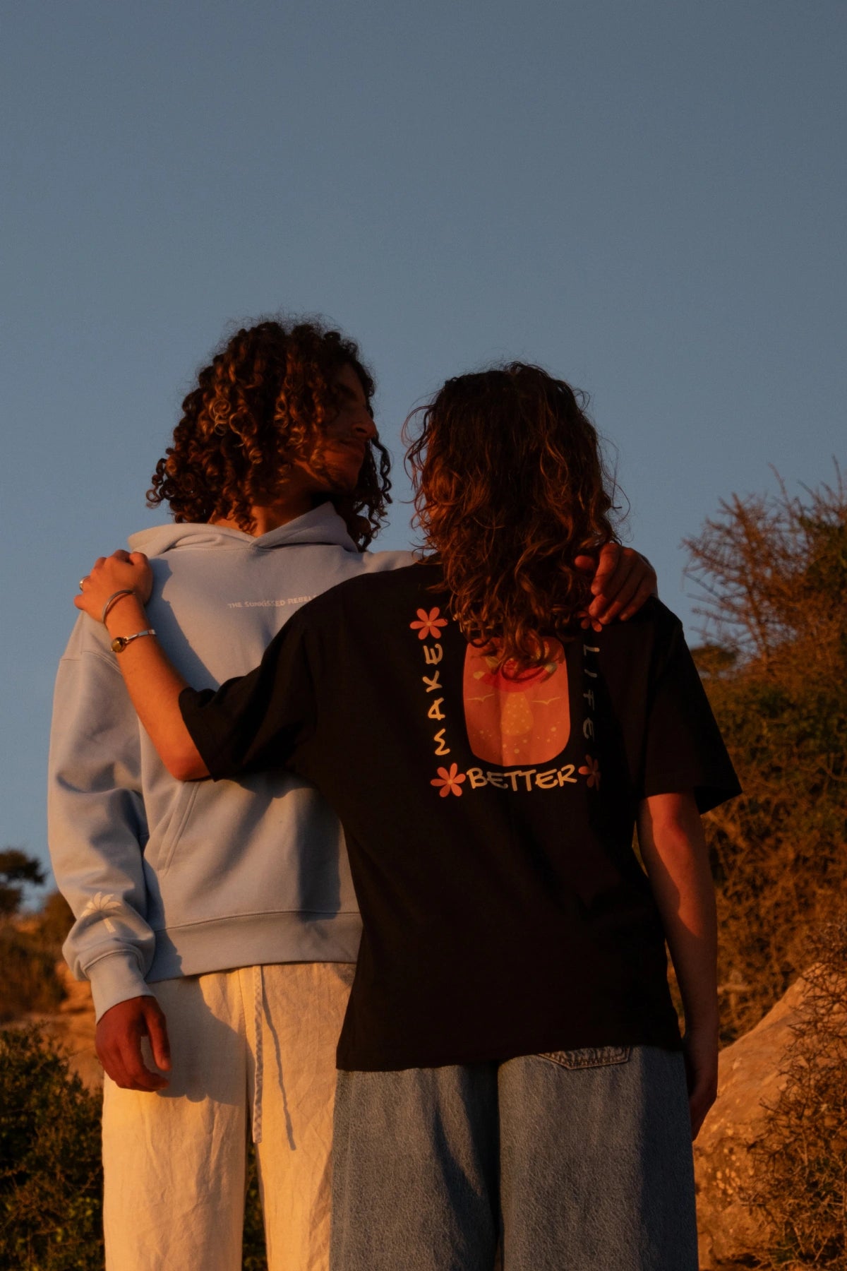 Zwei Models in nachhaltiger Surfwear bei Sonnenuntergang, mit Fokus auf den Backprint des schwarzen Good Life Light Tee in einer natürlichen Outdoor-Kulisse.