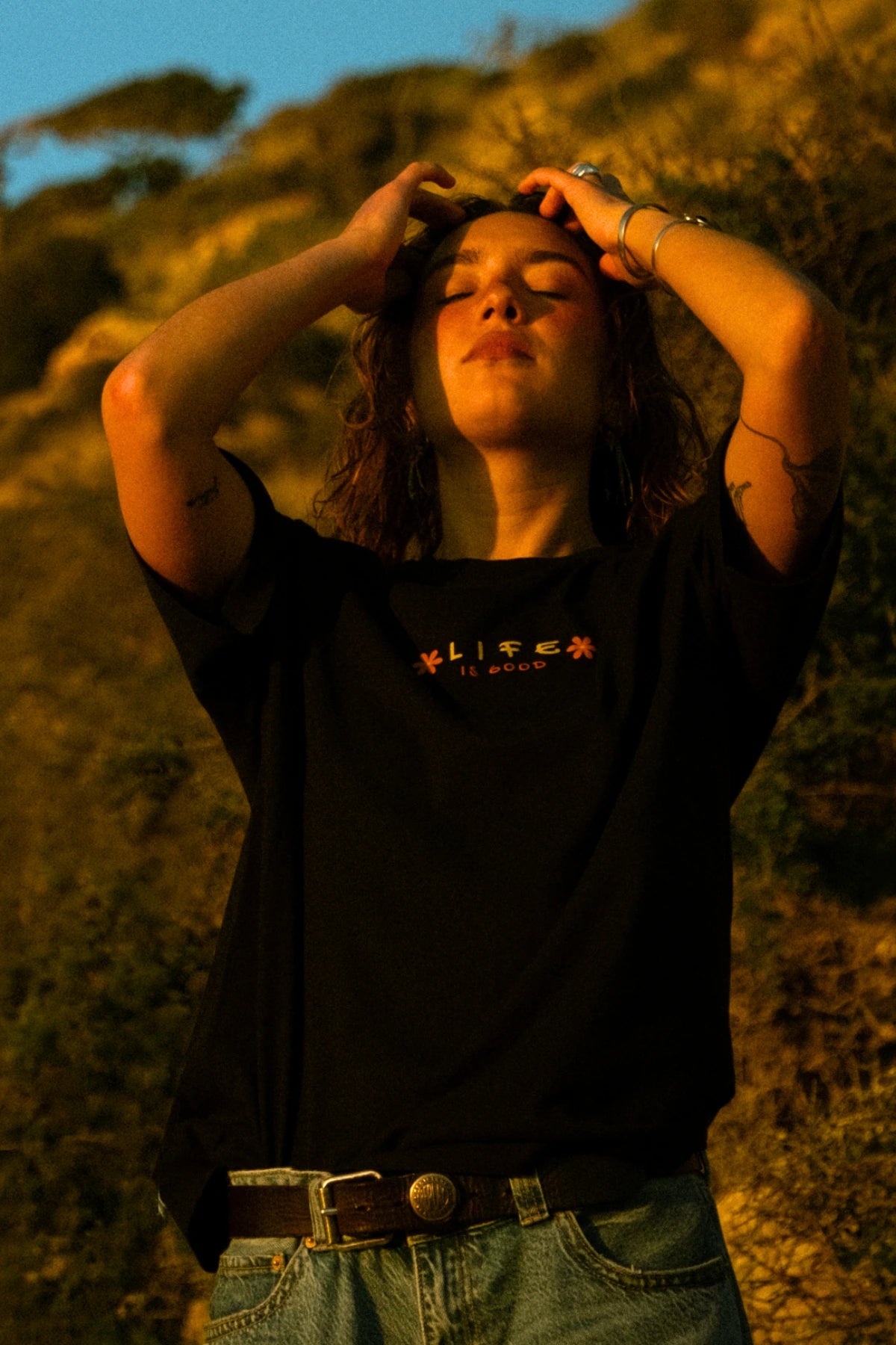 Weibliches Model trägt das schwarze Good Life Light Tee bei goldenem Sonnenuntergang in natürlicher Küstenlandschaft und verkörpert einen entspannten Beach-Lifestyle.