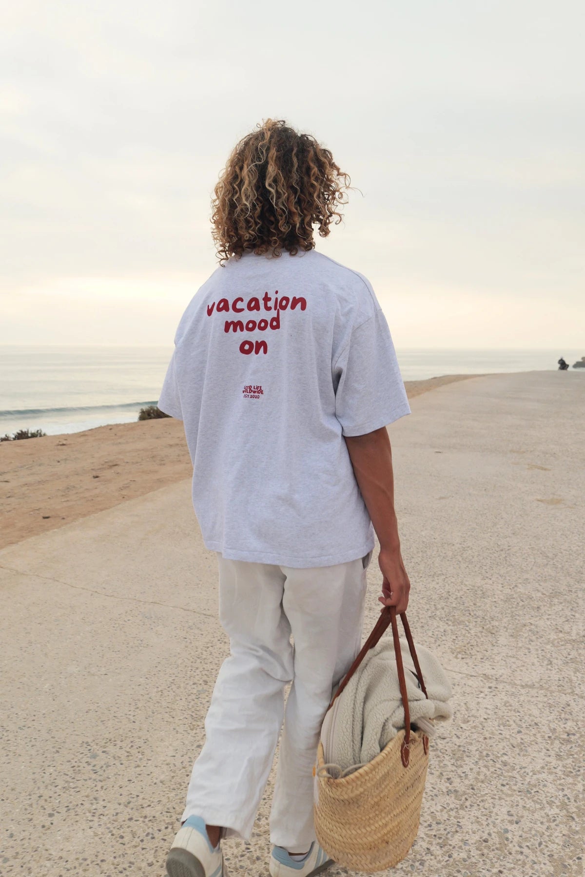 Model von The Sunkissed Rebel geht eine Küstenpromenade entlang und trägt das Goodlife T-Shirt in Cool Grey mit rotem Backprint “Vacation Mood On”.