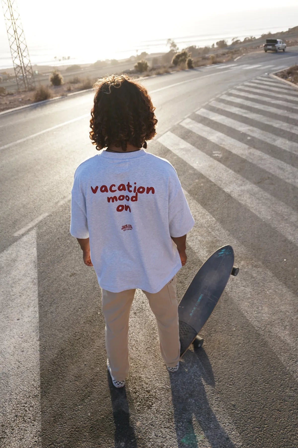 Nahaufnahme eines Models von The Sunkissed Rebel in der Sonne auf einer leeren Straße, trägt das Goodlife T-Shirt in Cool Grey und hält ein Skateboard.