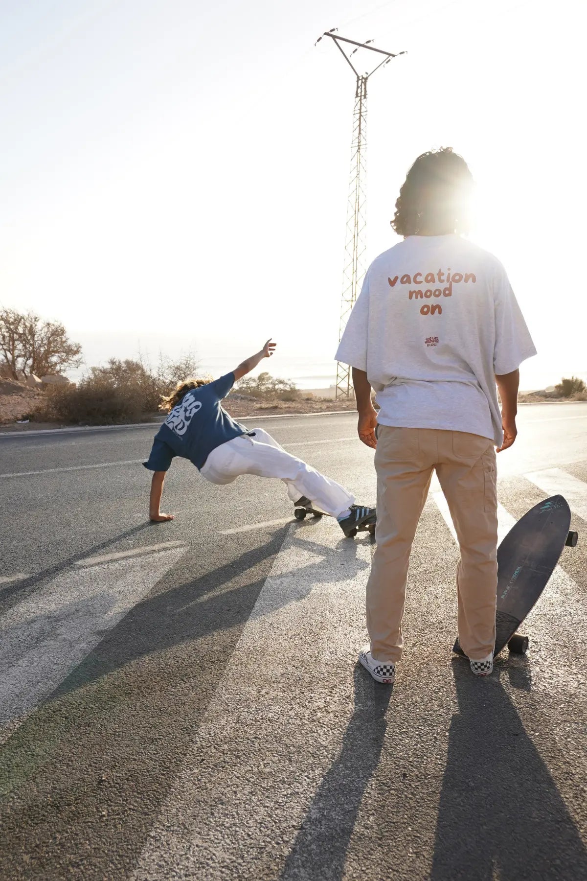 Model von The Sunkissed Rebel steht mit Skateboard auf einer sonnigen Küstenstraße, trägt das Goodlife T-Shirt in Cool Grey während ein anderes Model auf einem Longboard dynamisch vorbeifährt.