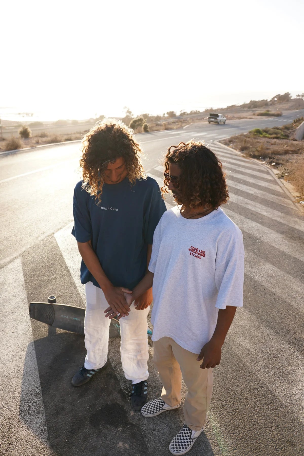 Zwei Models von The Sunkissed Rebel stehen auf einer Straße in der Abendsonne, beide tragen Streetwear und eines davon das Goodlife T-Shirt in Cool Grey, Longboard liegt daneben.