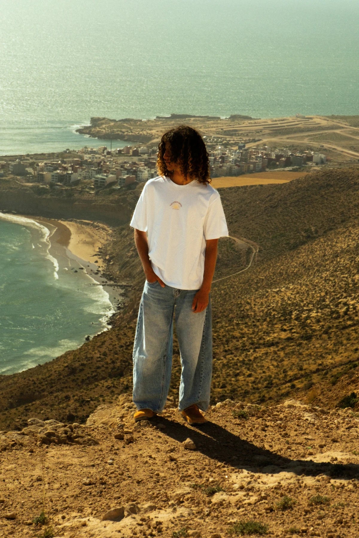 Männliches Model im weißen Iconic Light T-Shirt Pacific Chill von The Sunkissed Rebels bei warmem Sonnenlicht mit Ozeanpanorama im Hintergrund.