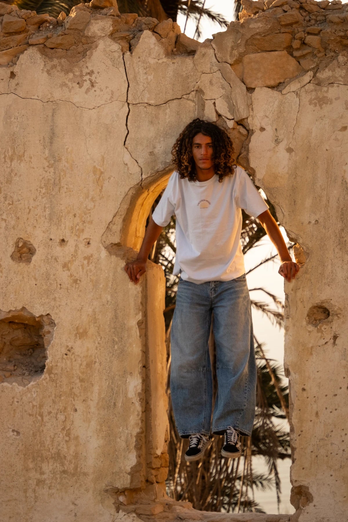Männliches Model trägt das weiße Iconic Light T-Shirt Pacific Chill von The Sunkissed Rebels vor einer historischen Ruine mit Palmen und warmem Sunset-Licht in tropischer Küstenkulisse.