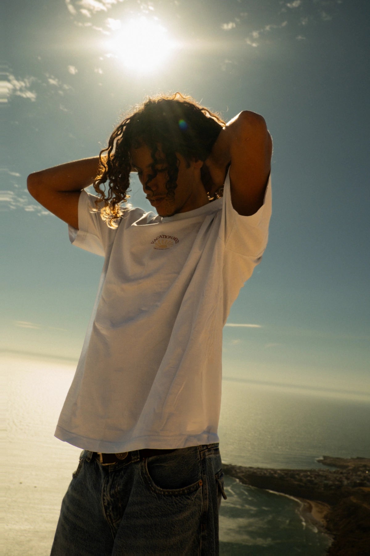 Männliches Model trägt das weiße Iconic Light T-Shirt Pacific Chill von The Sunkissed Rebels vor beeindruckender Küstenlandschaft mit Meerblick und Klippen.