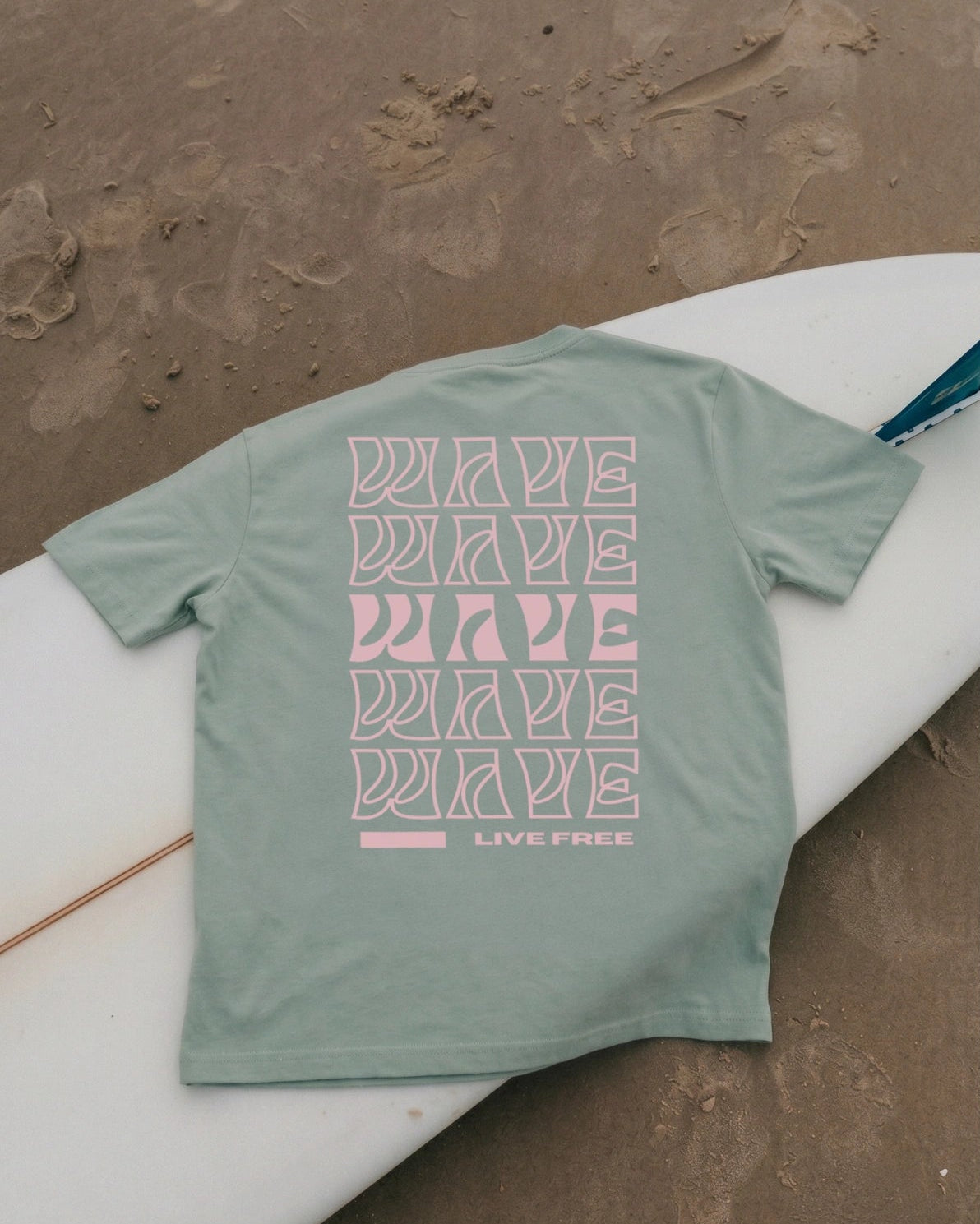 Aloegrünes Live Free Oversized T-Shirt von The Sunkissed Rebels auf Surfboard am Strand fotografiert