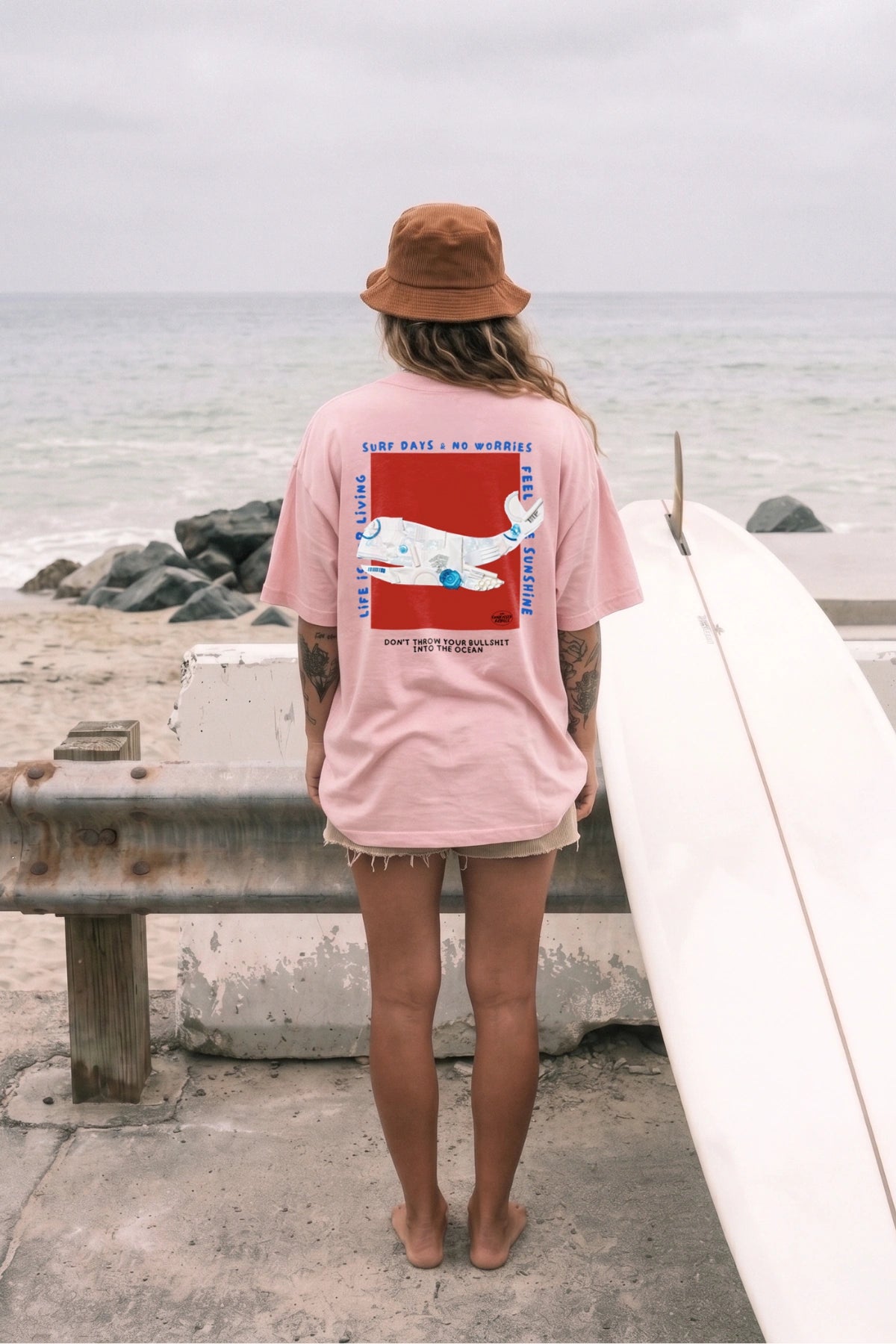 Rückansicht des No Bullshit Whale Boxy Oversized T-Shirts in Pink Joy mit großem Backprint neben einem Surfboard am Strand.