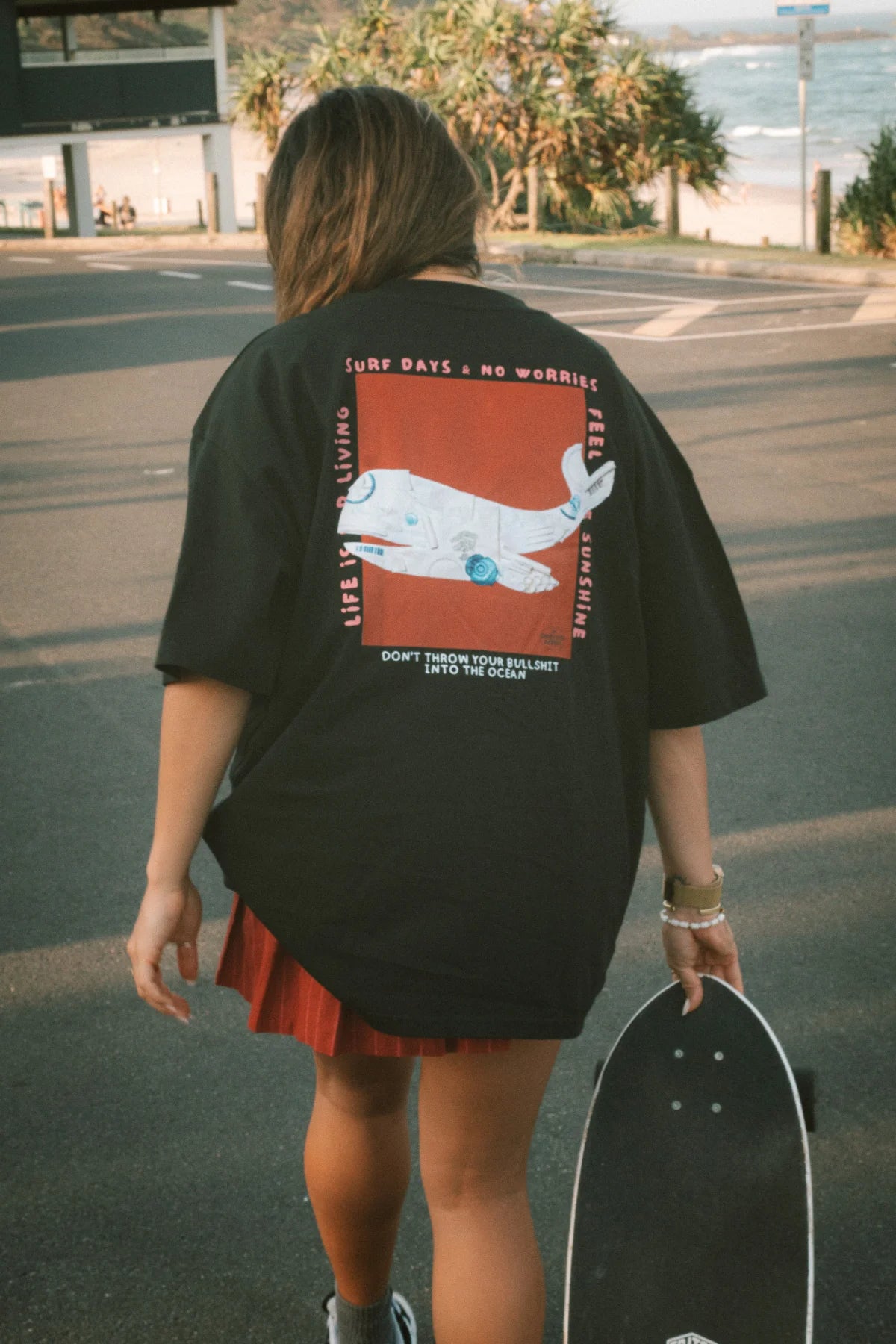 Skaterin mit dem No Bullshit Whale T-Shirt in Schwarz und rotem Rock vor dem Ozean – Casual Beach Outfit.