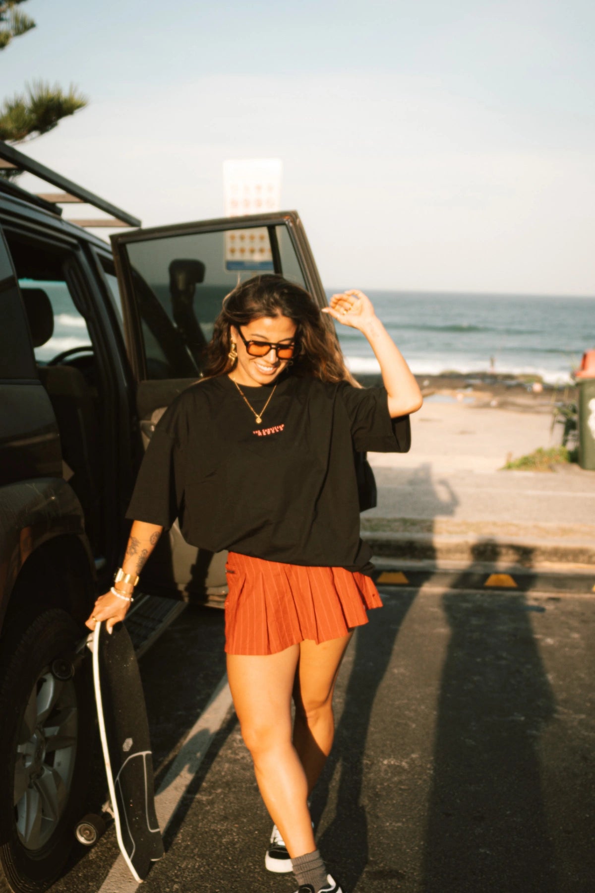 Frau mit Skateboard trägt das No Bullshit Whale T-Shirt in Schwarz am Strand – lässiger Surf-Look bei Sonnenuntergang.