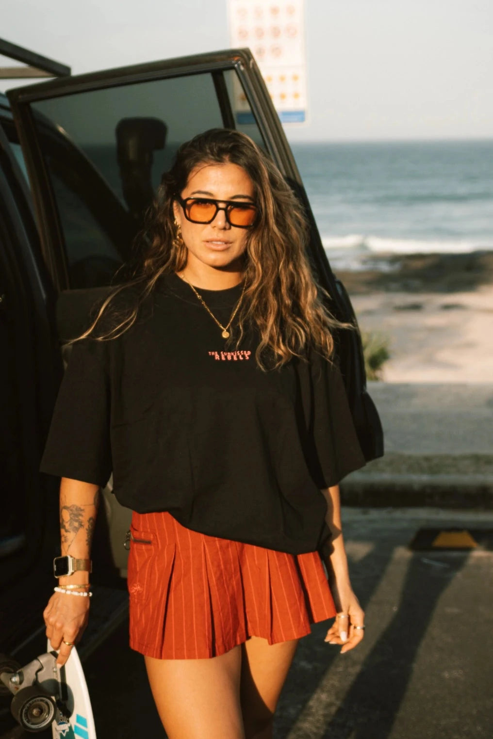 Lächelnde Frau trägt das No Bullshit Whale T-Shirt in Schwarz und roten Minirock vor dem Strand.