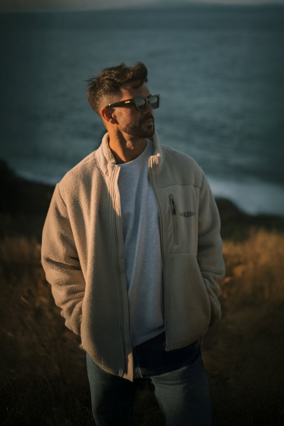 Mann steht am Meer mit  offener fleece Jacke in beige