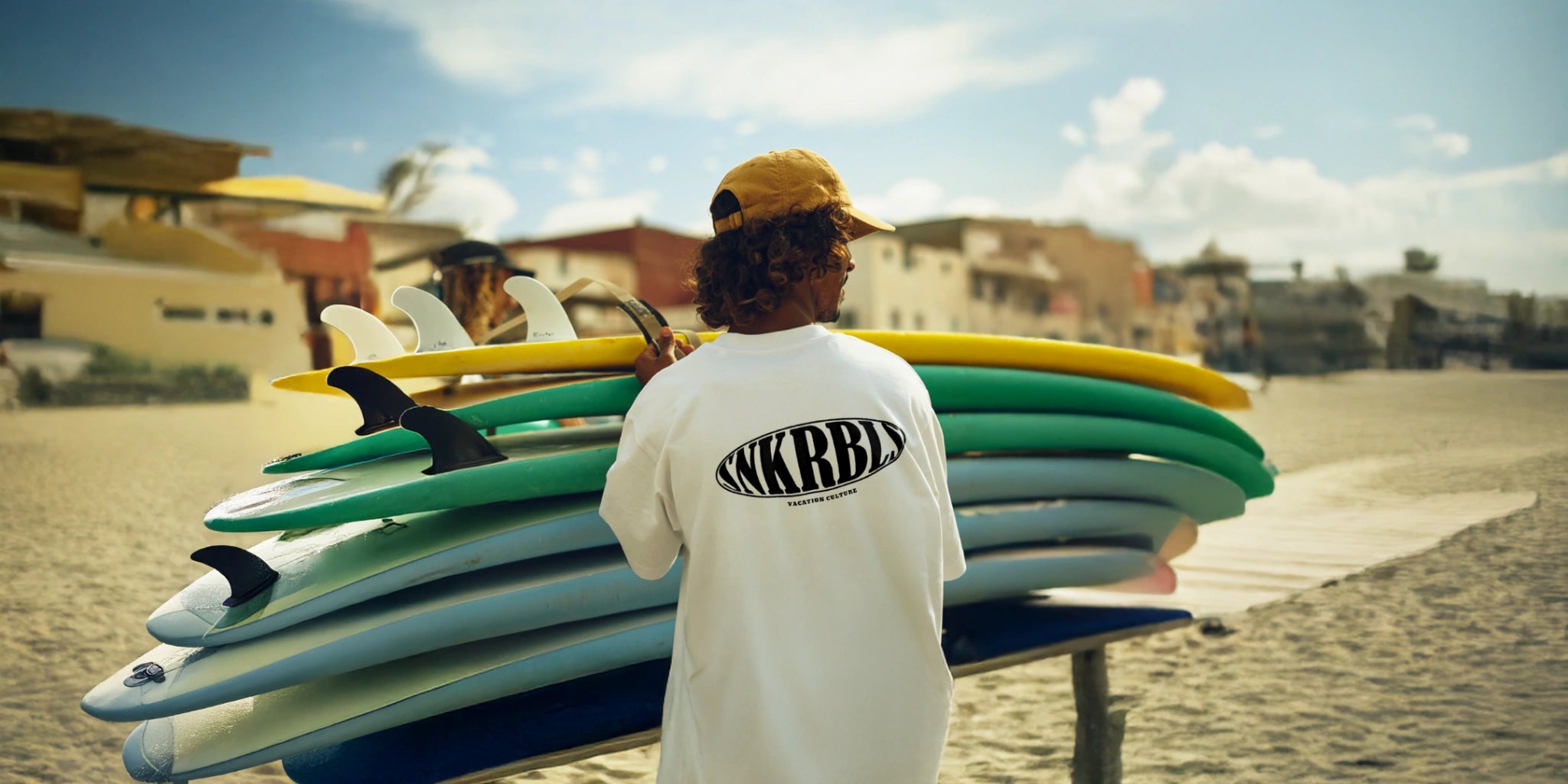 Person mit weißem Oversized T-Shirt mit Backprint vor gestapelten Surfbrettern am sonnigen Strand.