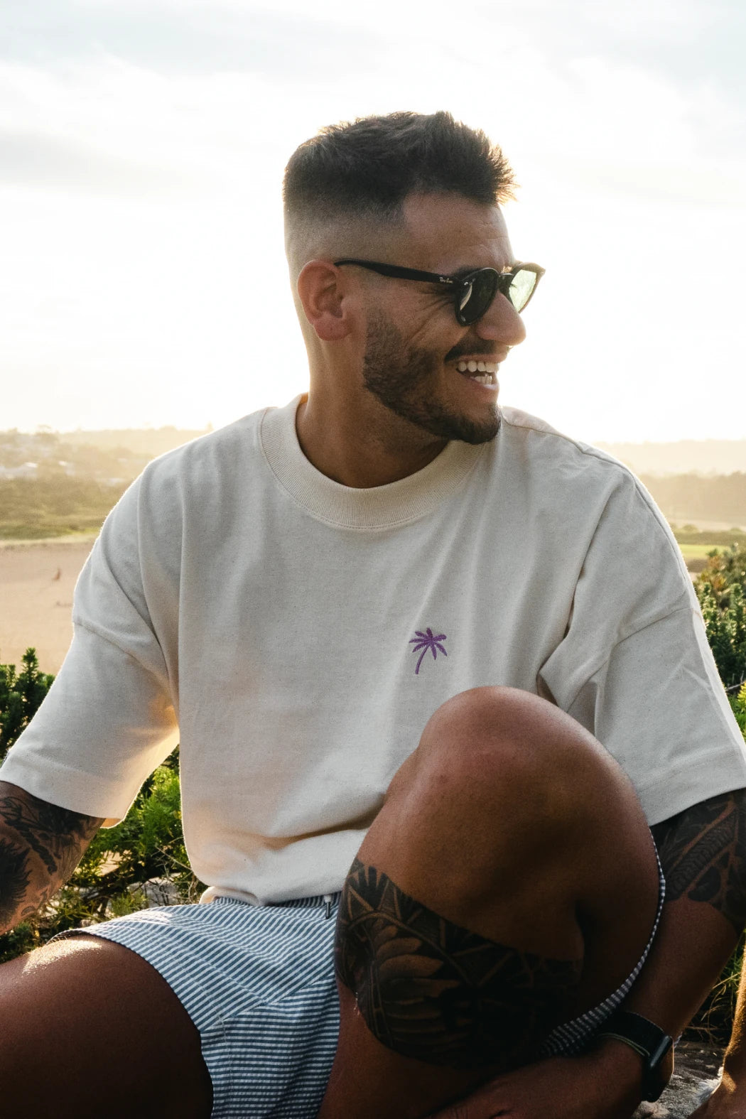 Lachender Mann im oversized T-Shirt in Natural Raw mit violettem Palmen-Print, bei Sonnenuntergang in der Natur sitzend.