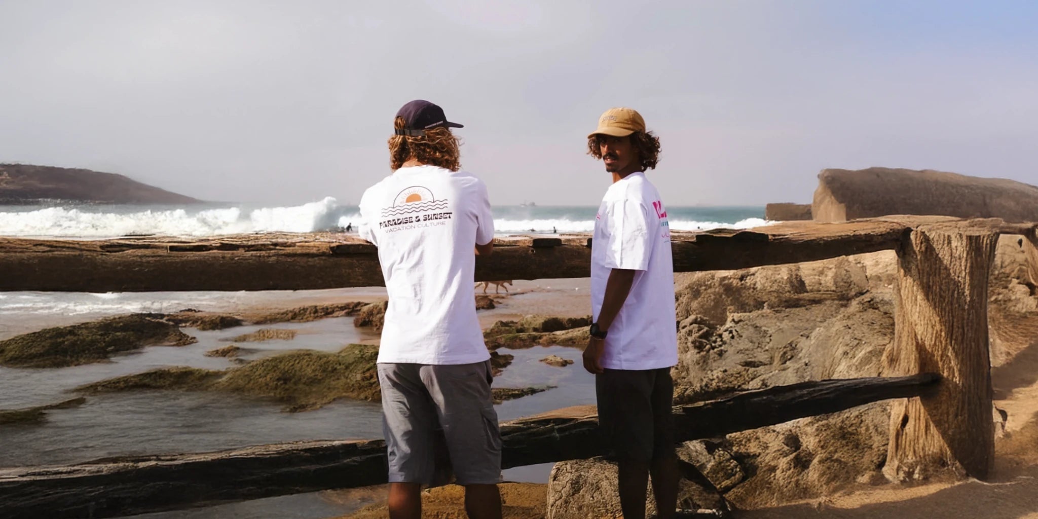 Zwei Männer in weißen T-Shirts mit Palmenprint stehen am Strand vor einer Holzabsperrung und blicken aufs Meer.