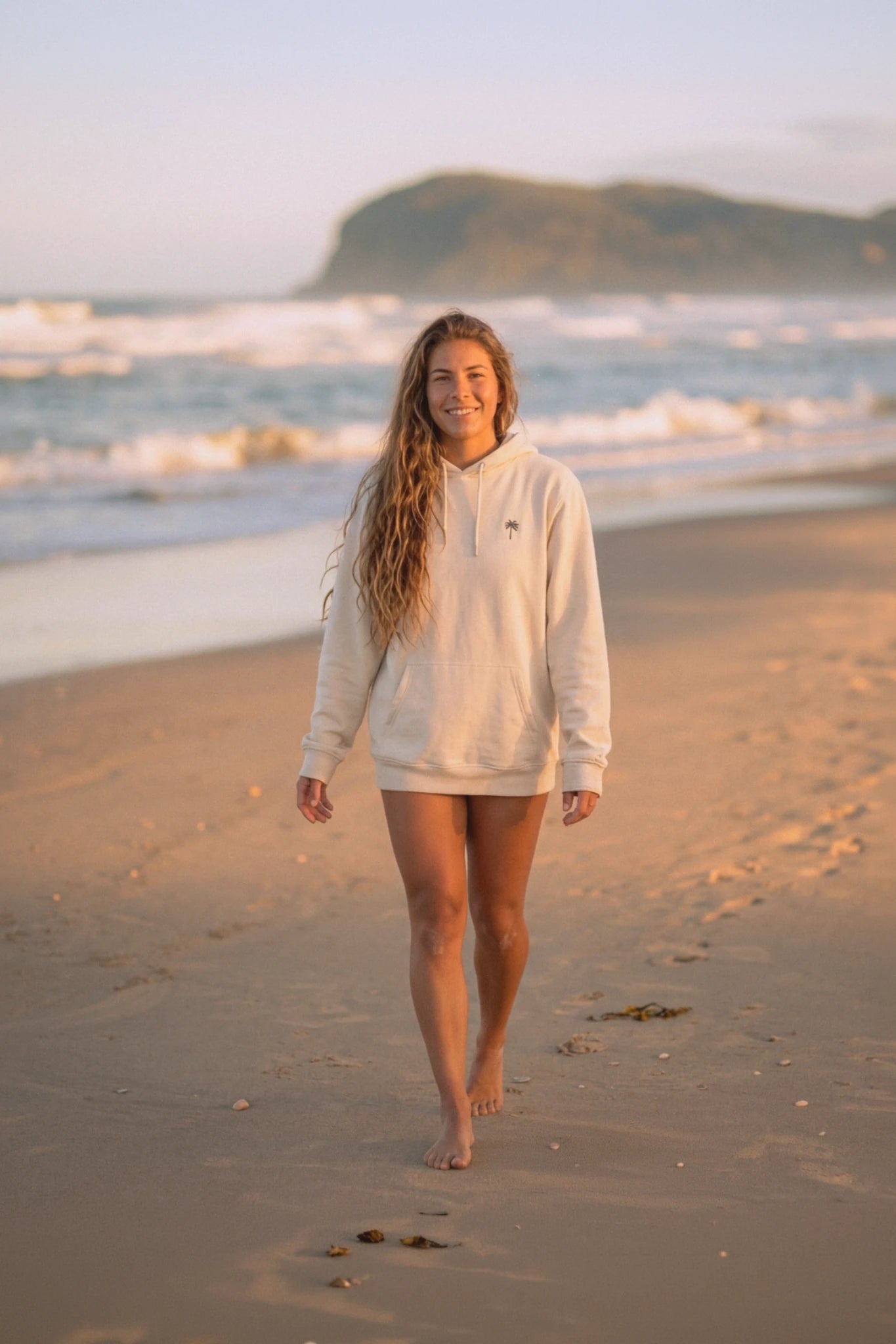 Frau läuft barfuß am Strand bei Sonnenuntergang und trägt Palm Tree Hoodie in Natural Raw – minimalistischer nachhaltiger Hoodie im Beach Style