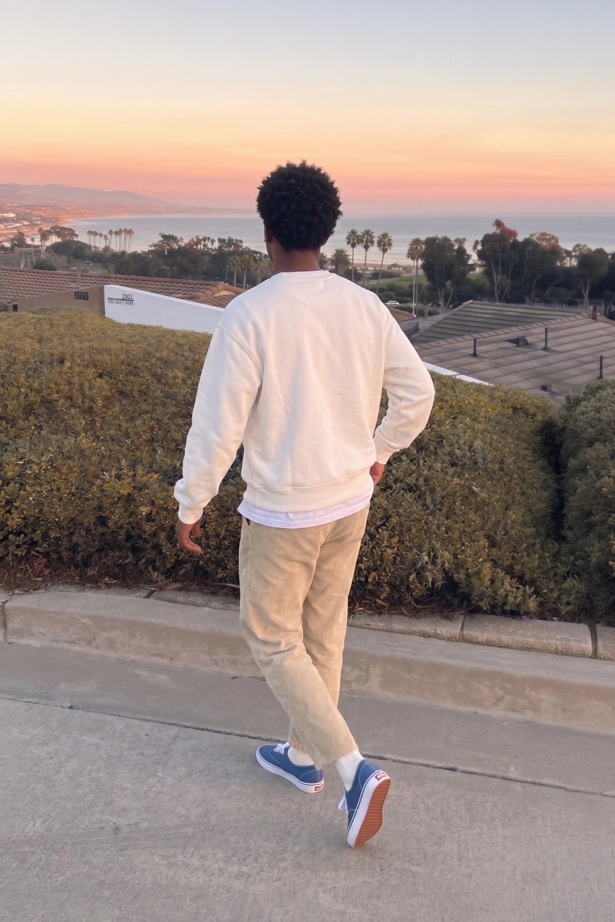 Rückenansicht eines Models im cremefarbenen Palm Tree Oversized Sweater mit Meerblick bei Sonnenuntergang