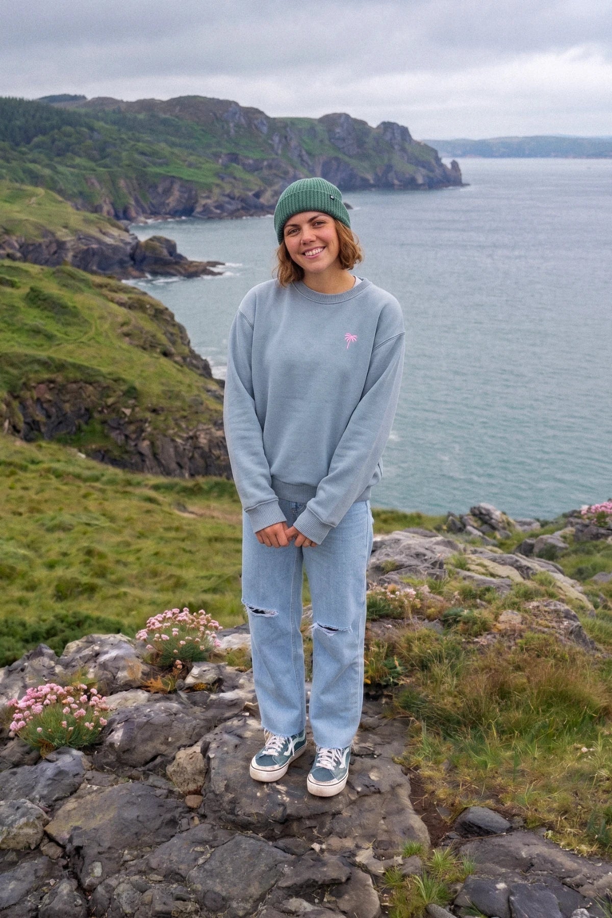 Frau trägt den Pink Palm Oversized Vintage Sweater in Serene Blue an einer grünen Küstenlandschaft mit Blick auf das Meer.