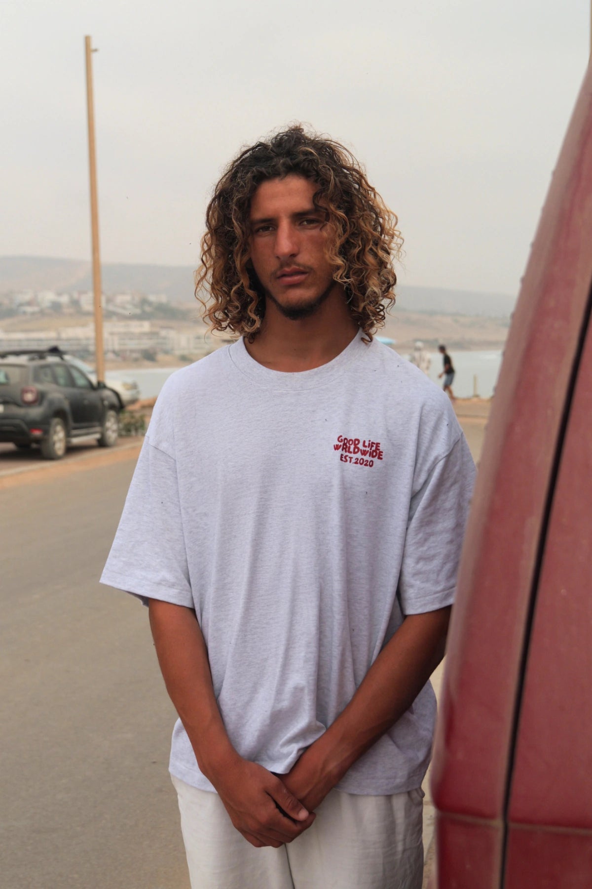 Model von The Sunkissed Rebel trägt das Goodlife T-Shirt in Cool Grey und steht entspannt an einer Straße mit Küstenblick.