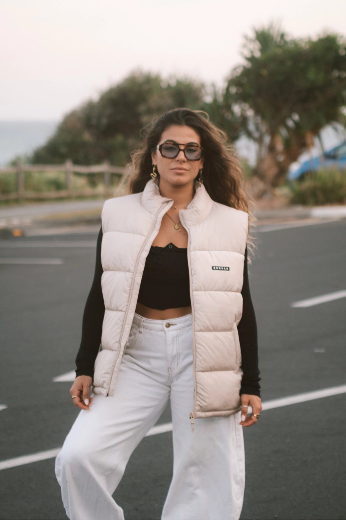 Streetstyle-Look – Frau mit der Rebels Puffer Outdoor Vest Beige von The Sunkissed Rebels steht auf einem Parkplatz und schaut in Kamera
