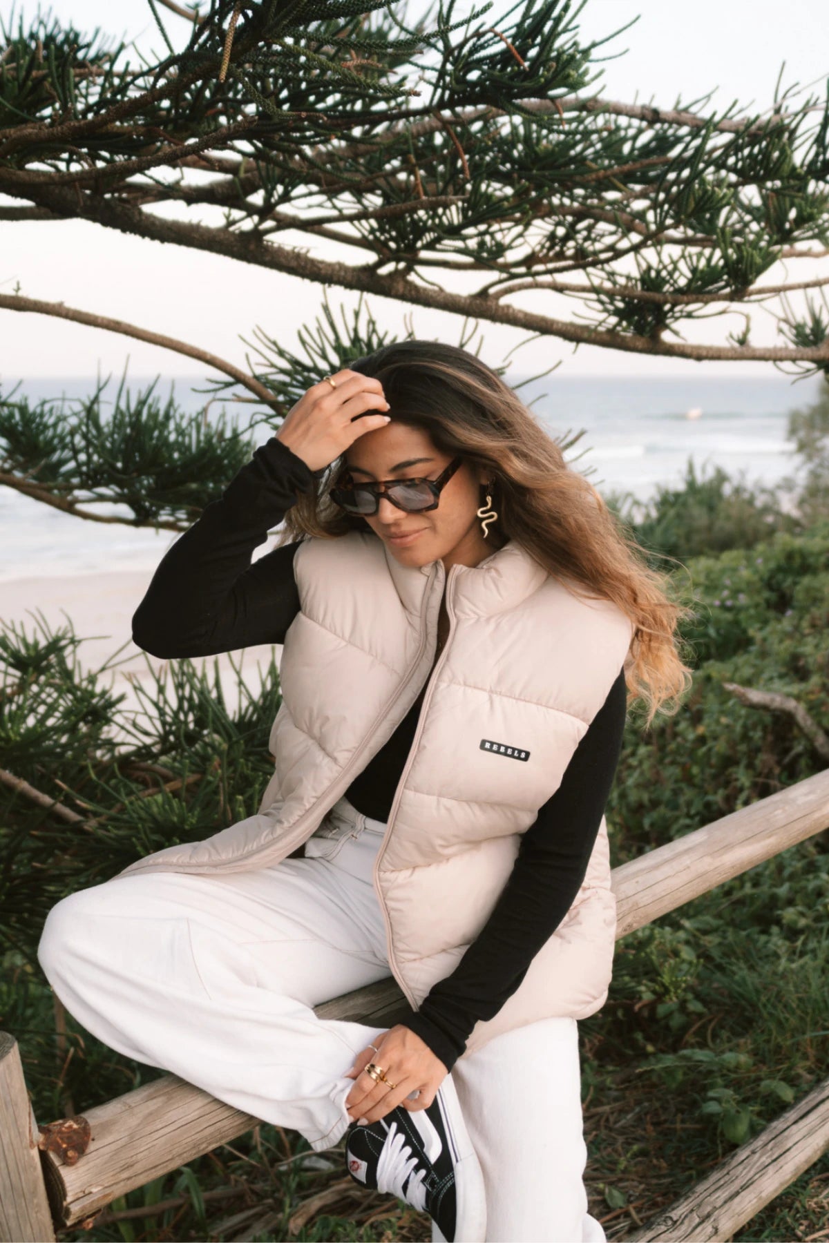 Streetstyle-Look – Frau mit der Rebels Puffer Outdoor Vest Beige von The Sunkissed Rebels sitzt auf Holzbalken am Strand
