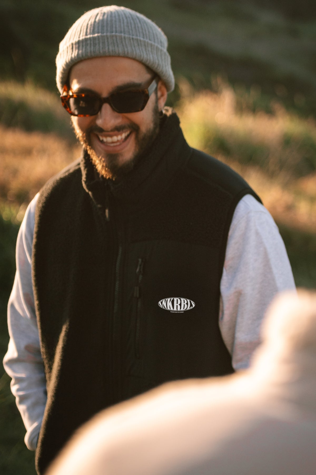 Mann steht auf Hügel und lacht bei Sonnenuntergang und trägt schwarze outdoor Weste 