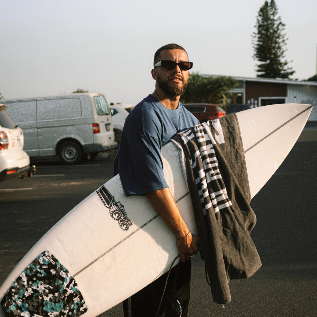 Mann mit blauem „SURF CLUB“-T-Shirt trägt Surfbrett und Handtuch, bereit für die Wellen am Strand.