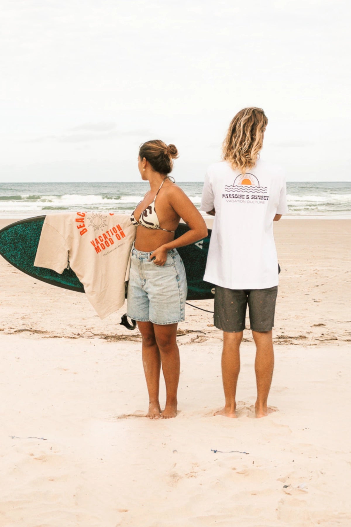 Rückansicht von Mann im weißen Paradise-&-Sunset-Shirt und Frau mit Surfbrett am Strand