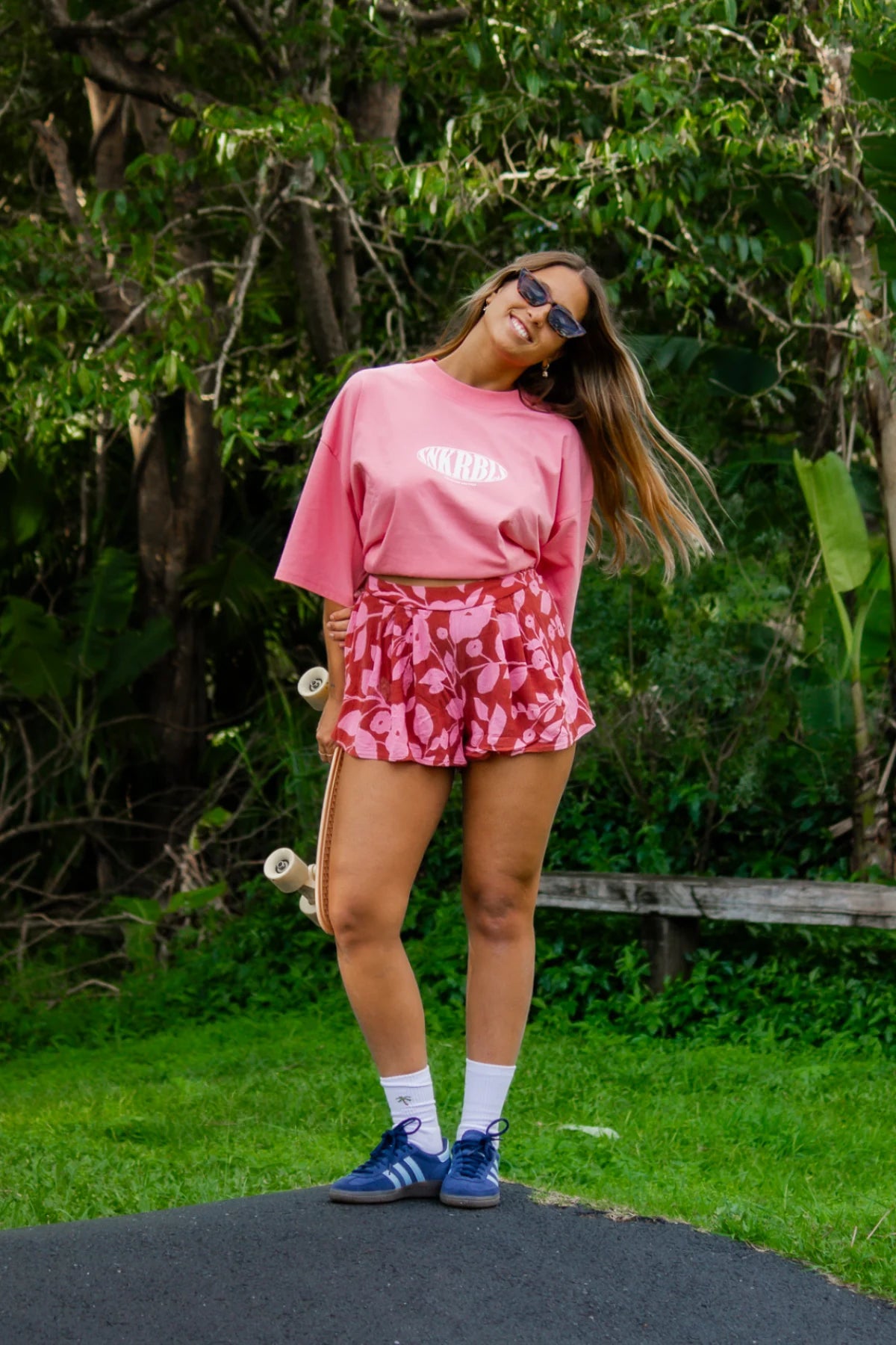 Glückliche Frau im SNKRBLS T-Shirt Pink Joy mit floralem Rock und Skateboard in der Natur.