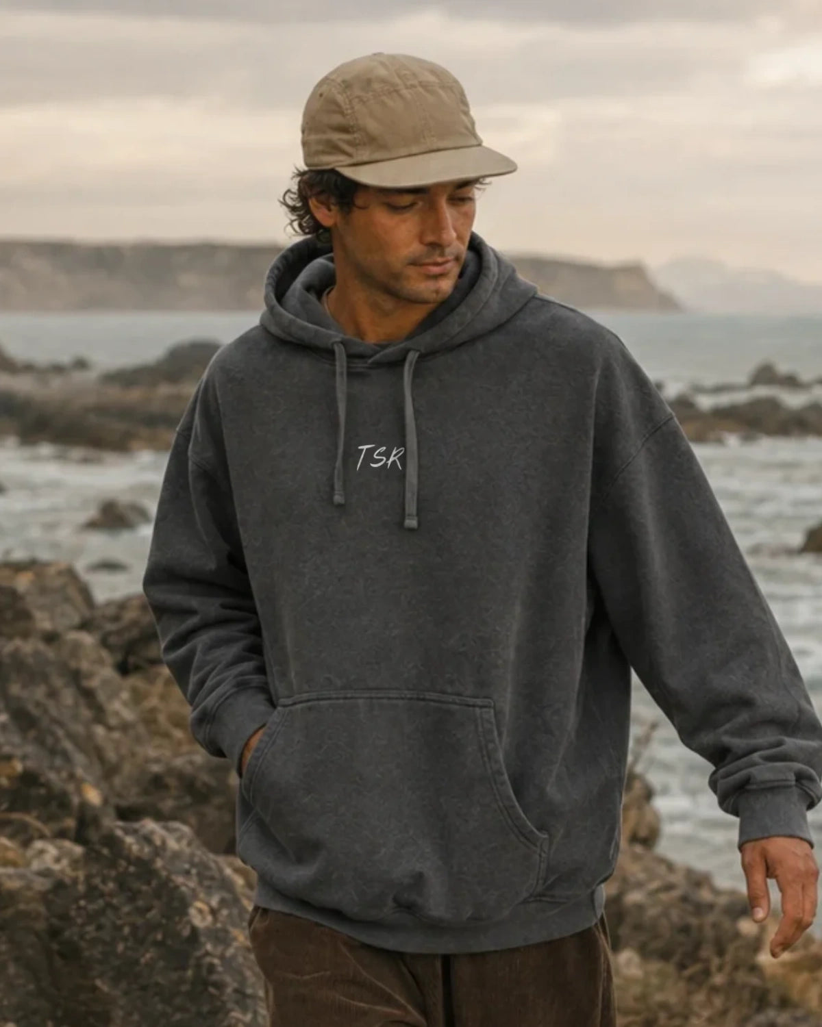 Mann in Vintage Anthracite Hoodie mit dezentem TSR-Schriftzug, beiger Cap und lässiger Passform vor einer felsigen Küstenlandschaft.