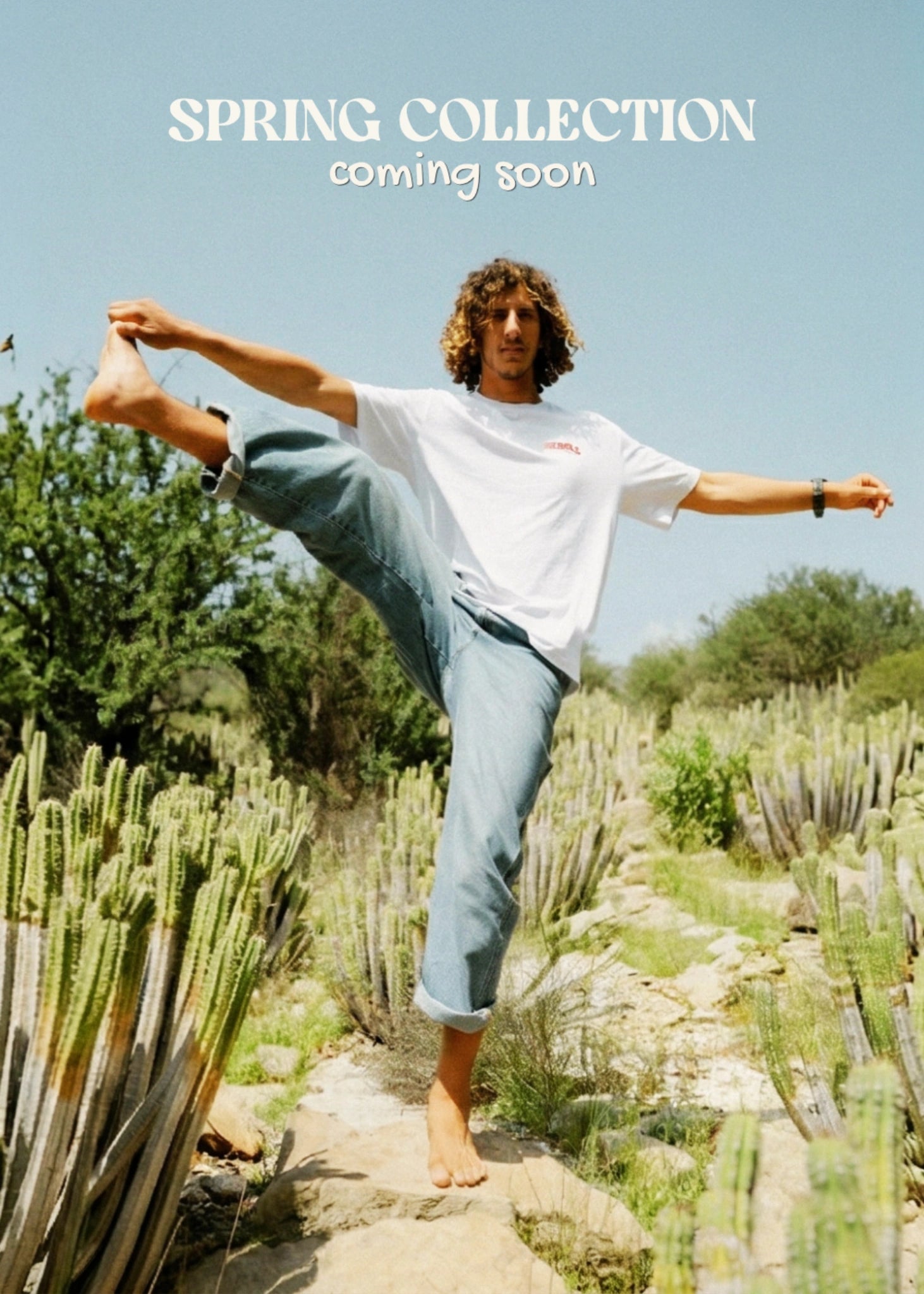 Model trägt weißes T-Shirt aus der Spring Collection und macht Stretch-Pose in Wüstenlandschaft mit Kakteen