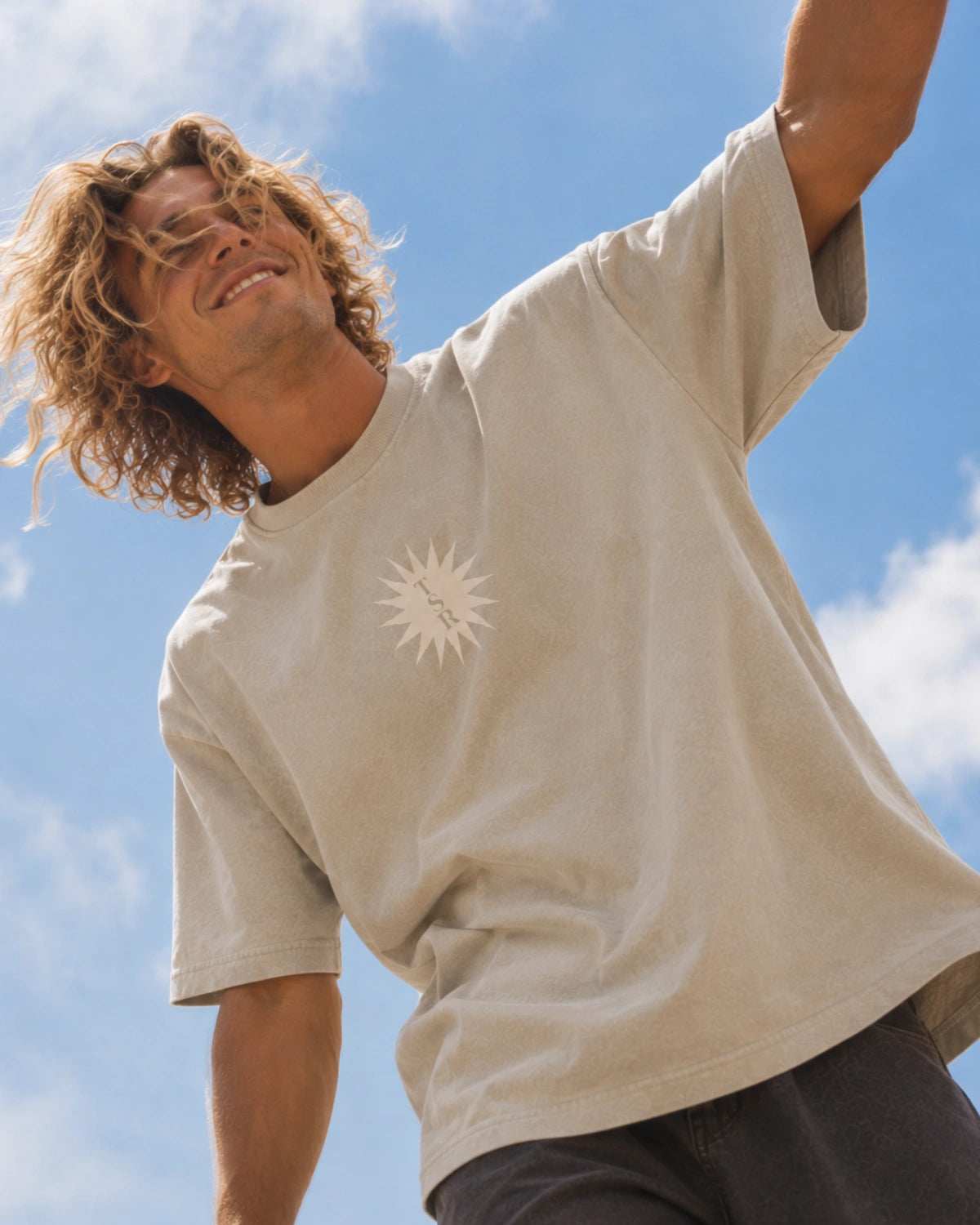 Nahaufnahme des Sunburst Boxy Tees in Stone mit TSR-Sonnenprint auf der Brust, getragen von einem Mann vor blauem Himmel.