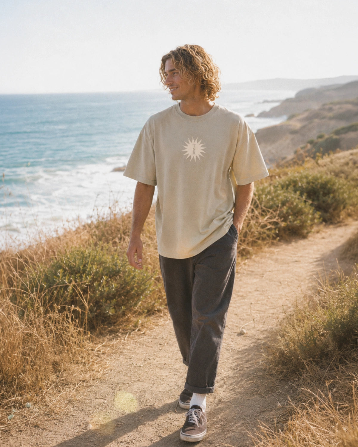 Mann trägt das beige Sunburst Boxy Tee mit lockerem Oversized Fit und weißem Sonnenprint auf einem Küstenweg am Meer.