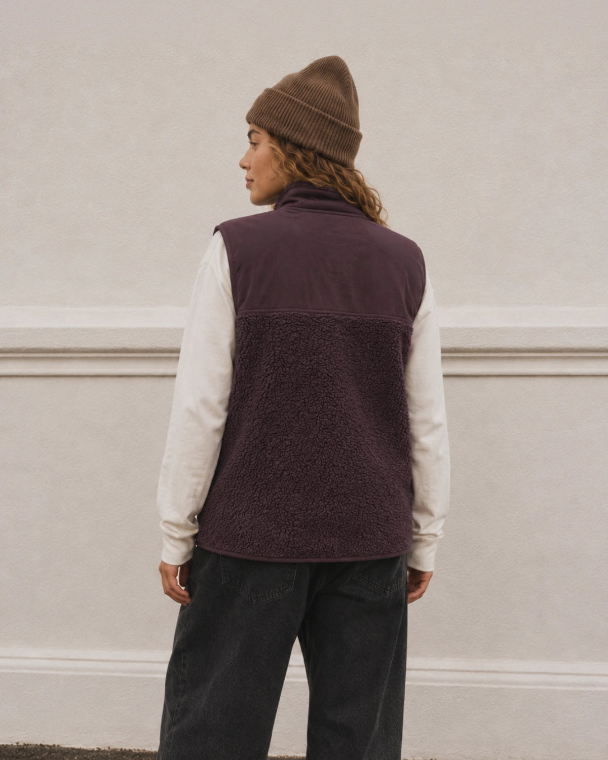 Frau trägt die Sundown Fleece Vest in Burgundy von The Sunkissed Rebels über einem weißen Longsleeve, kombiniert mit brauner Beanie und schwarzer Jeans.