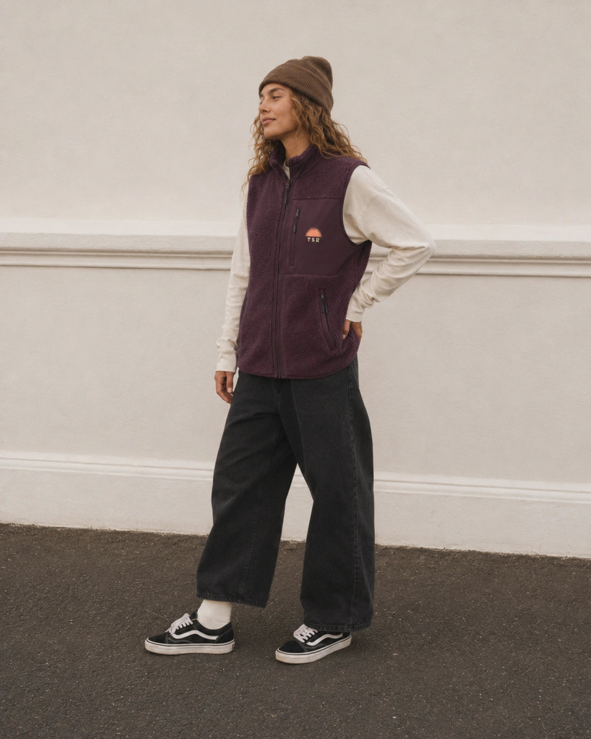 Frau trägt die burgunderrote Sundown Fleeceweste mit weißem Longsleeve, weiter schwarzer Jeans und Sneakers. Lässiger Unisex-Look.