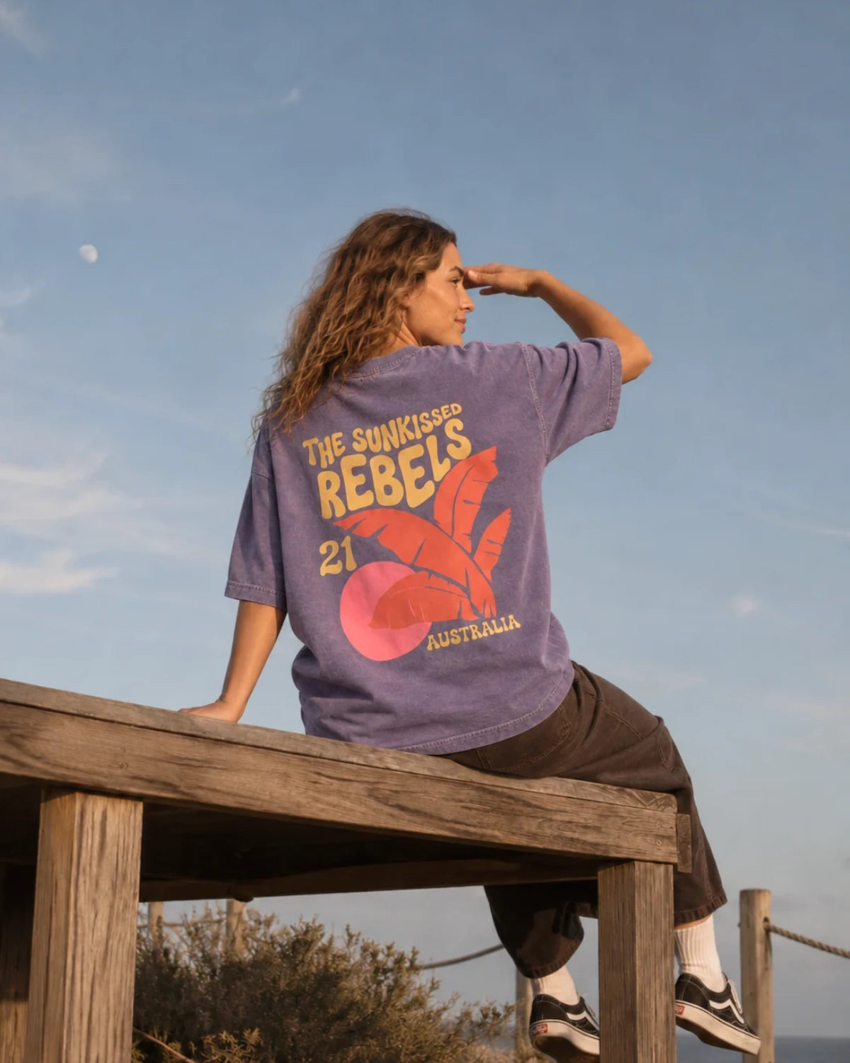 Frau sitzt auf einer Holzplattform am Meer und trägt das Sunkissed Rebels Heavy Tee in Vintage Violet mit farbigem Palmen-Backprint.