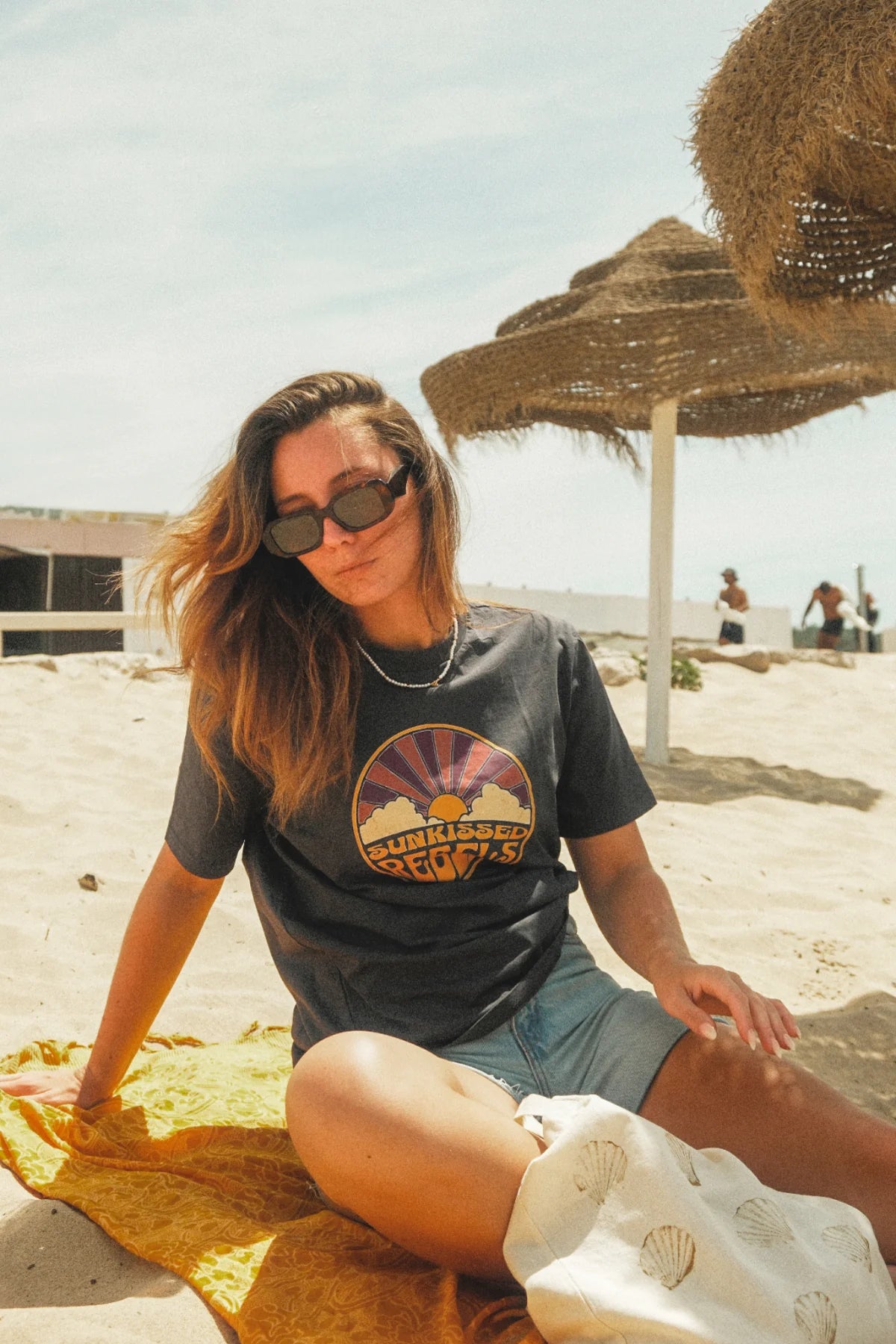 Frau mit India Ink Grey T-Shirt und Sonnenbrille am sonnigen Strand mit Sonnenschirm.
