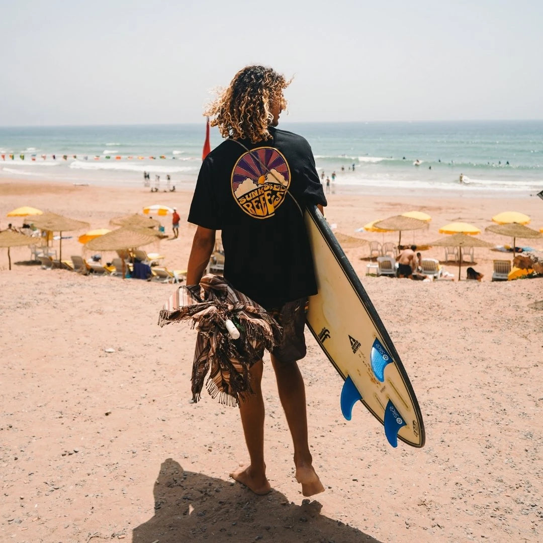 Surfer mit Sunkissed Rebels Shirt und Surfbrett am sonnigen Strand