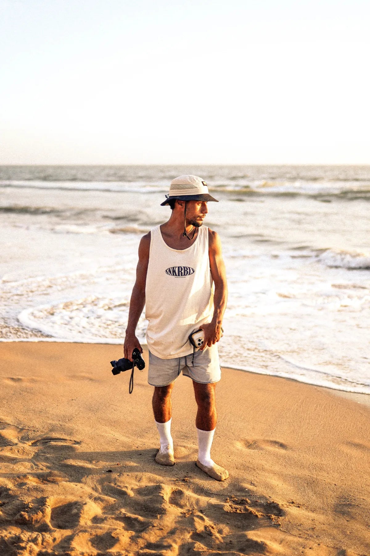 Mann am Strand mit Kamera im SNKRBLS Tank Top Natural Raw – lässiger Sunset-Look mit Bucket Hat