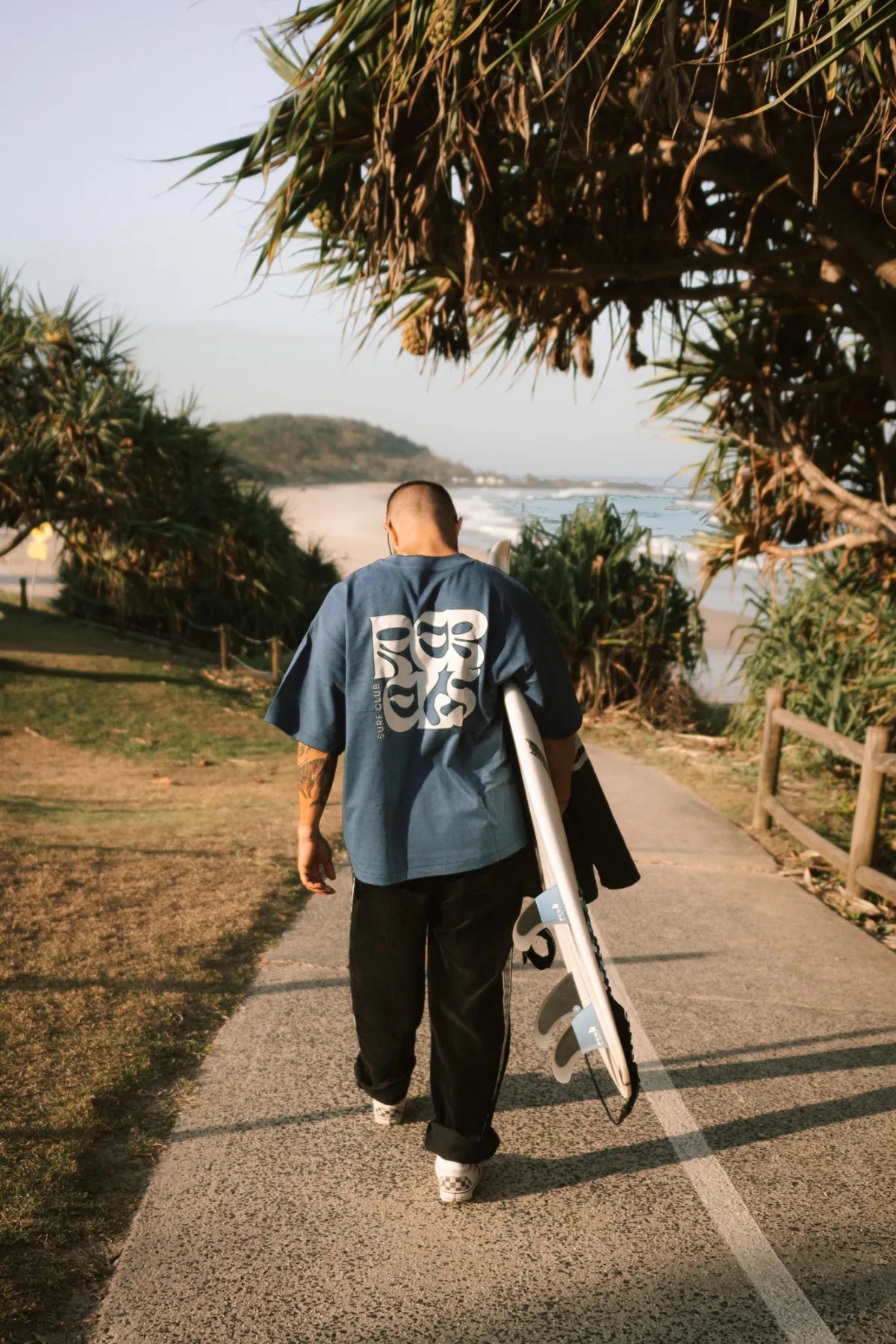 Mann mit Surfbrett beim Spaziergang am Strand im Surf Club Heavy T-Shirt Deep Blue von The Sunkissed Rebels – lässiger Sommerstil.