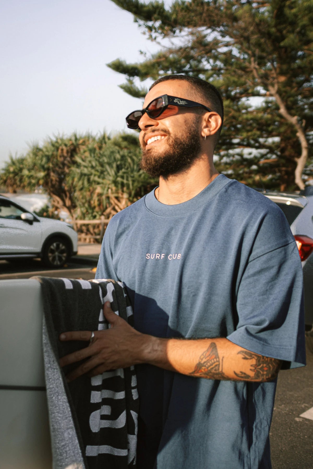 Entspannter Look am Surfspot – Surf Club Heavy T-Shirt Deep Blue von The Sunkissed Rebels mit Surfbrett, perfektes Outfit für Strandtage.