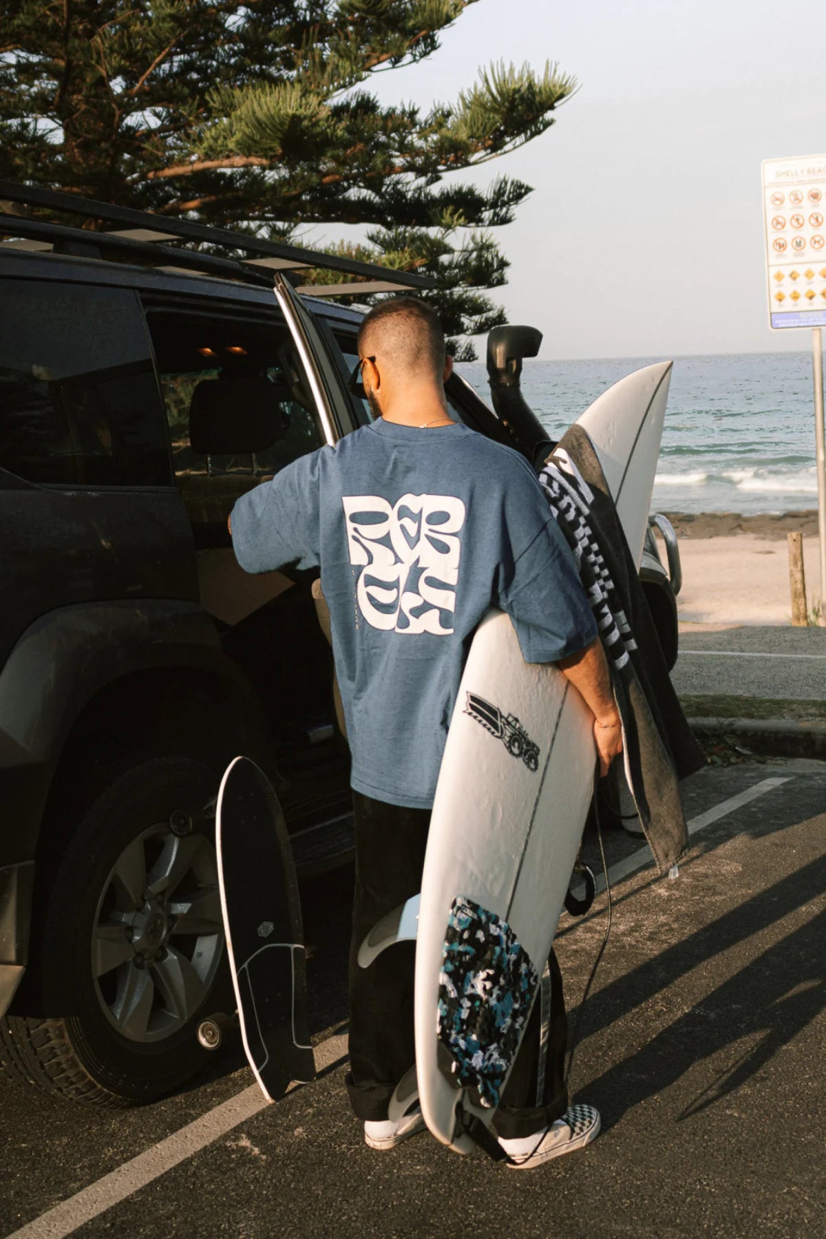 Surf Club Heavy T-Shirt in Deep Blue von The Sunkissed Rebels – Surfer lädt Board in den Van, authentischer Beach Lifestyle Moment.