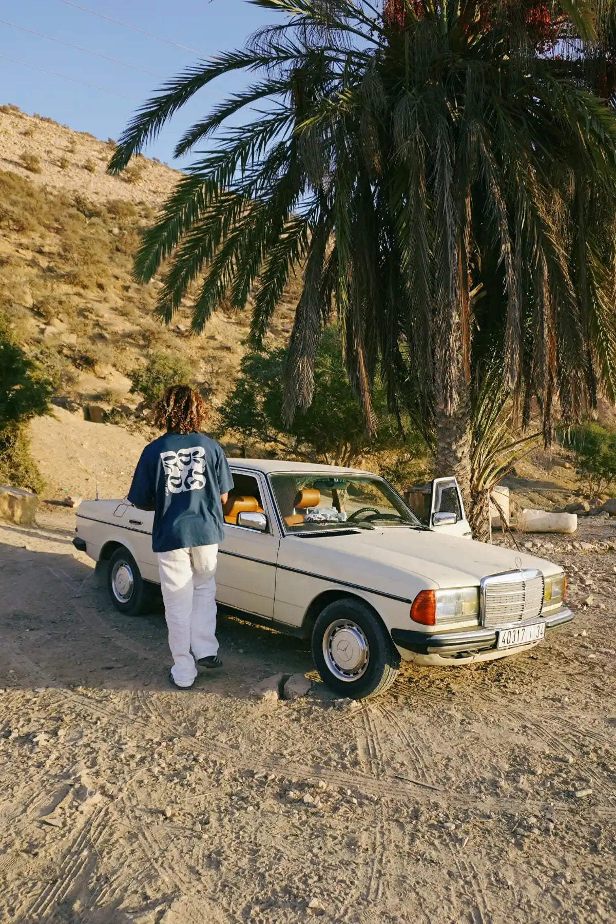 Model von The Sunkissed Rebel geht neben einem Oldtimer in einer Wüstenlandschaft und trägt das Surf Club T-Shirt in Deep Blue mit großem Rückenprint.
