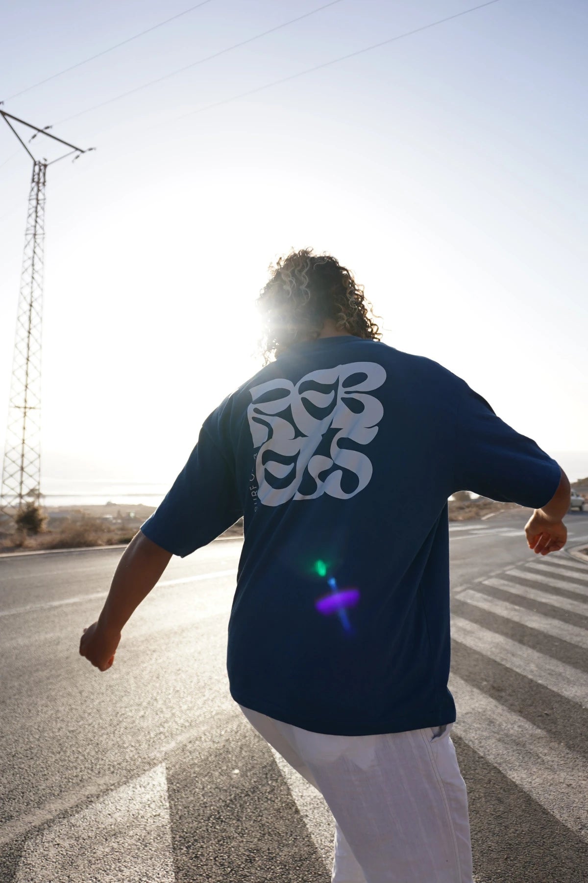 Model von The Sunkissed Rebel fährt dynamisch auf einem Skateboard eine kurvige Straße entlang und trägt das Surf Club T-Shirt in Deep Blue.